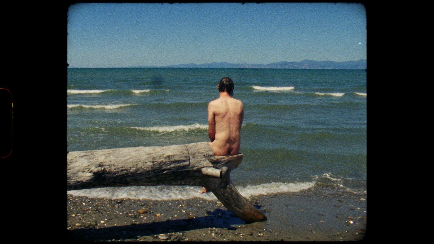 På stranden sitter en naken mann på en horisontal trestamme med ryggen til og ser ut mot havet.
