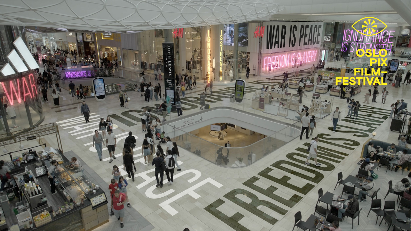Oversiktsbilde fra et moderne kjøpesenter med mange mennesker i bevegelse. Store neonskilt og gulvtekst viser slagord som «WAR IS PEACE», «FREEDOM IS SLAVERY» og «IGNORANCE IS STRENGTH». Butikker som H&M, Adidas og John Lewis er synlige i bakgrunnen.
