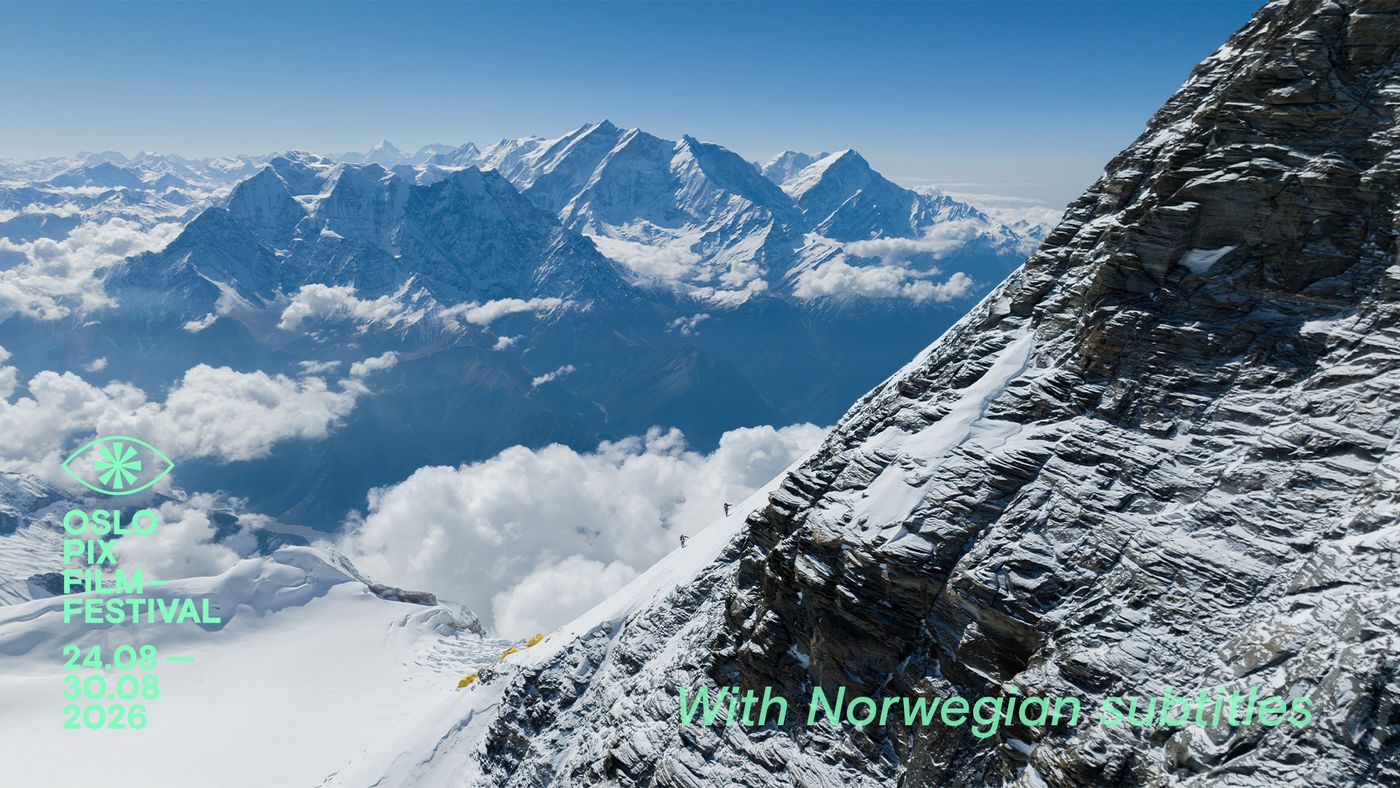 To personer går opp en bratt, snødekt fjellside. Flere snødekte fjelltopper og skyer i bakgrunnen.