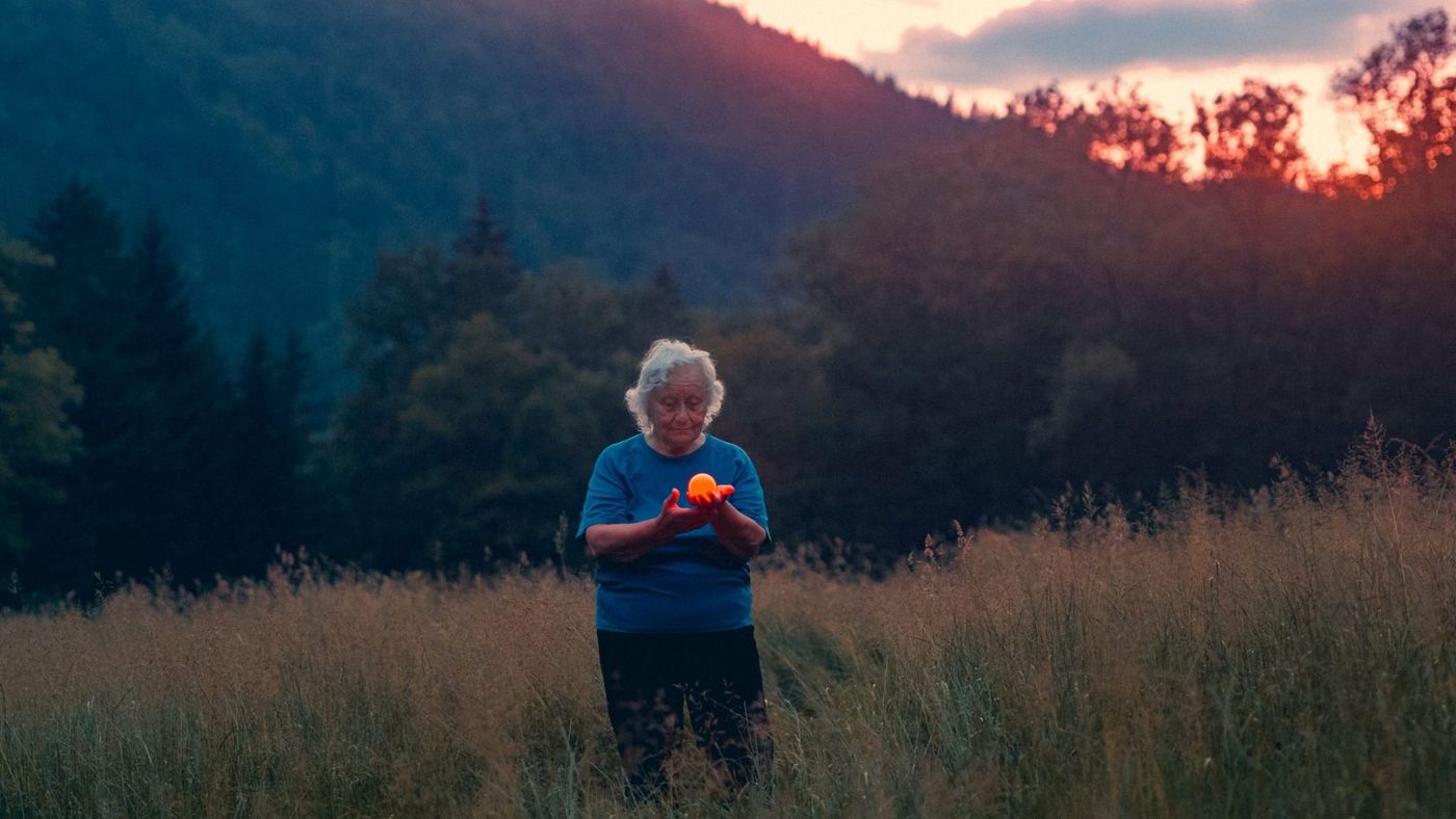 Bildet viser en eldre dame stående i en eng omringet av høye strå. Det er solnedgang og lyset skinner av rød- og blåtoner. I bakgrunnen står et fjell med trær. Kvinnen har hvitt krøllete kort hår og er kledt i blå t-skjorte og mørke bukser. Hun holder en orange lysende ball på størrelse med en tennisball.