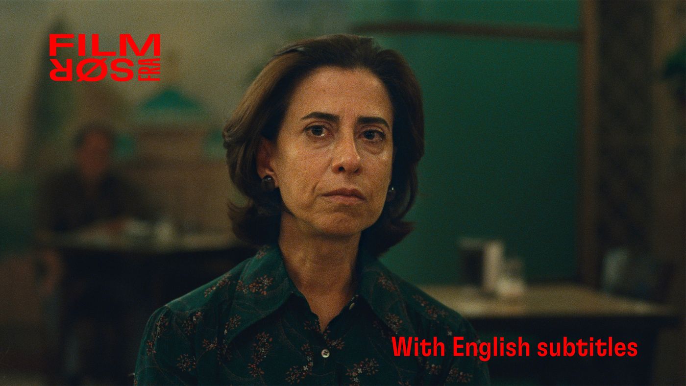 Kvinne med skulderlangt hår og grønn skjorte ser mot kamera. Uskarpt interiør med bord og stoler i bakgrunnen.