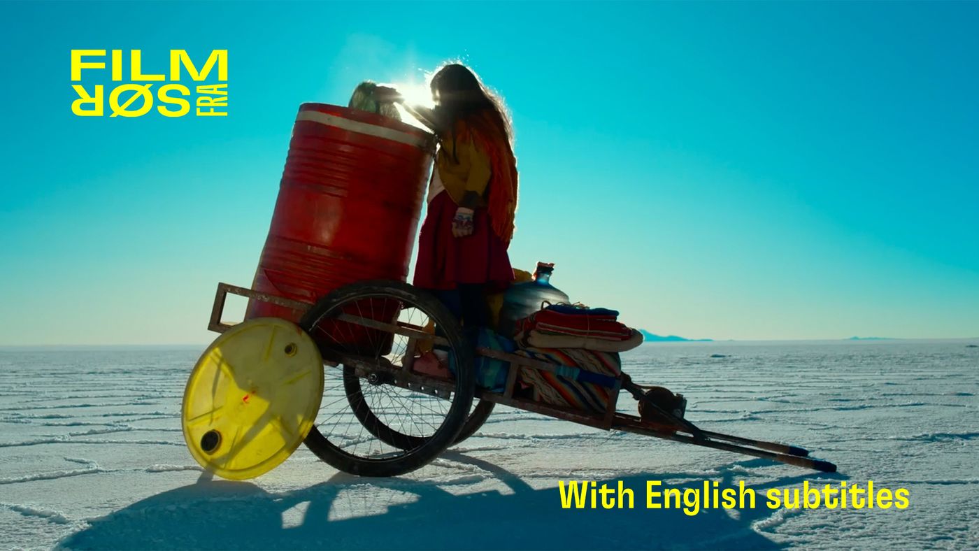 Person med langt hår står ved en tralle med store beholdere i et åpent, hvitt landskap under klar himmel.