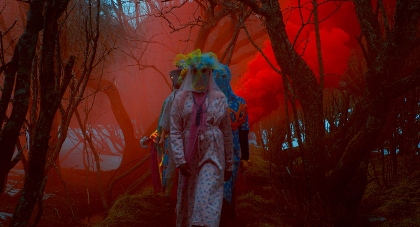 En gruppe kvinner i fargerike kostymer og tildekkede ansikt går igjennom skogen. De er omsluttet av rød røyk