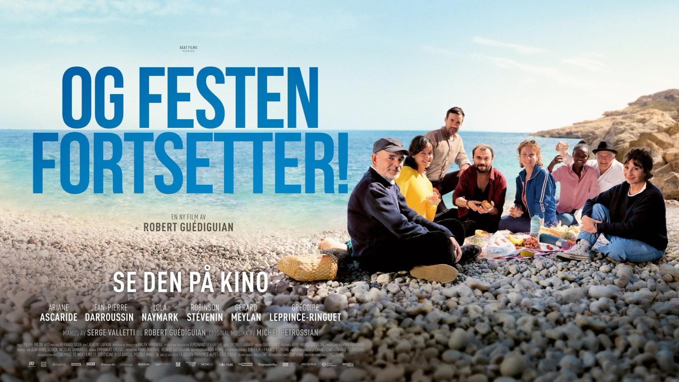 en gruppe mennesker som sitter på en steinete strand ved havet og nyter et måltid sammen, smilende og med drinker i hendene.