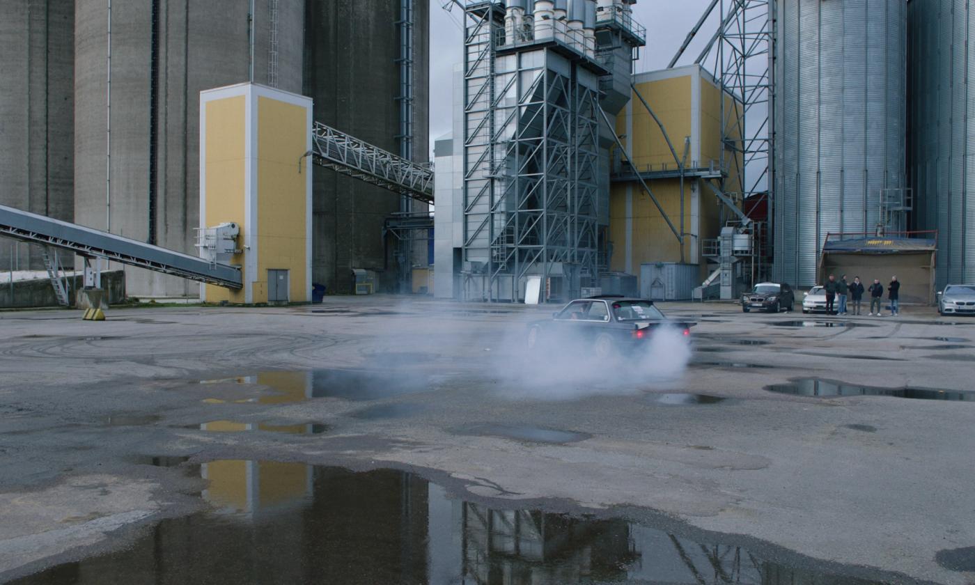En bil drifter på et industriområdet foran fem menn som ser på.