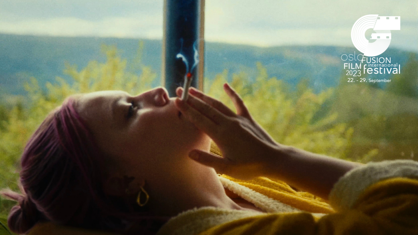 En ung kvinne ligger med en sigarett opp til munnen med natur utenfor vinduet ved siden av henne.