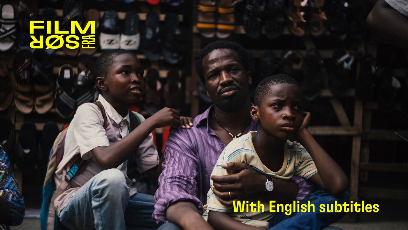 En mann sitter omgitt av sko med to gutter ved siden av seg, begge med skolesekker.