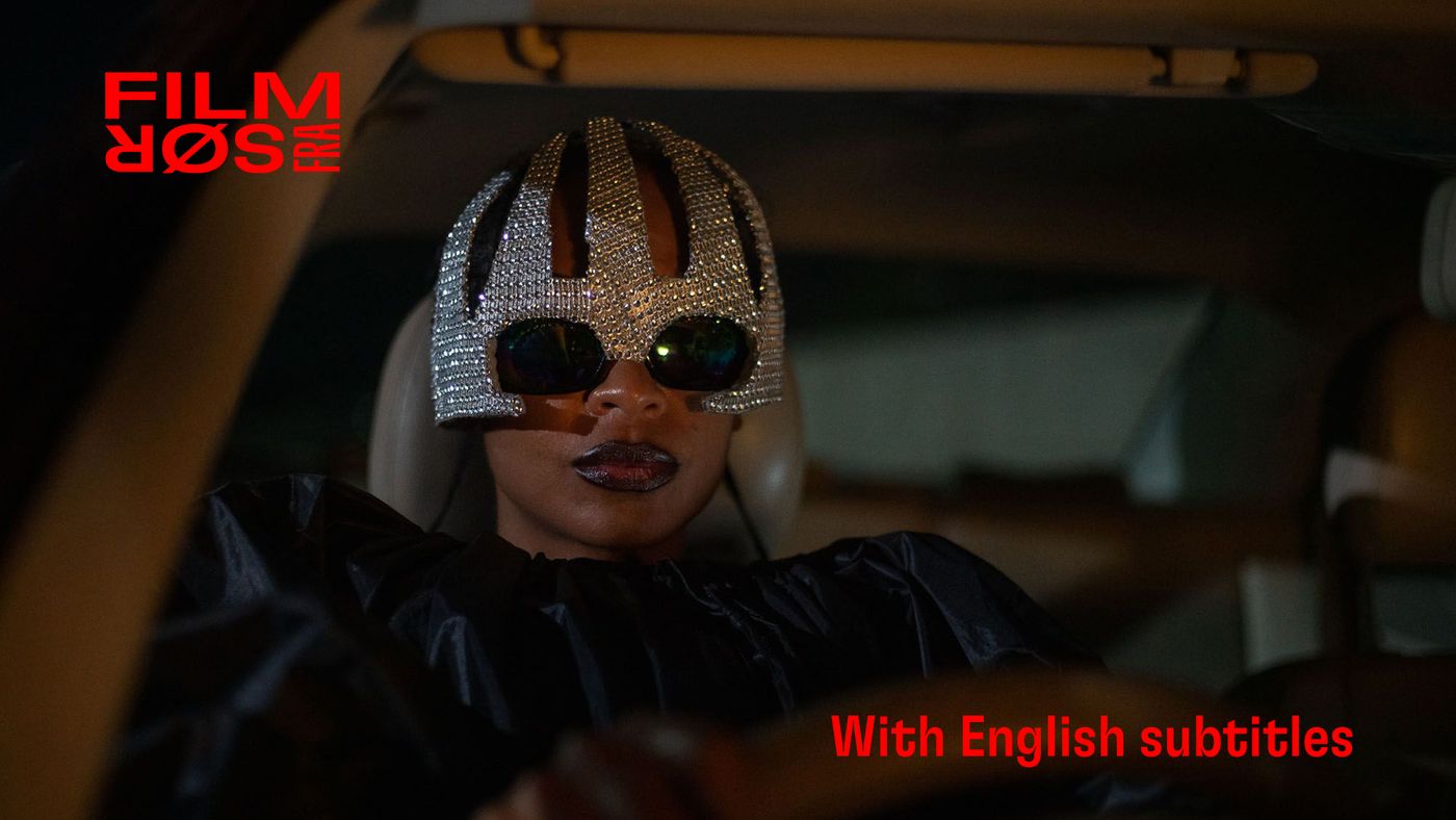 Kvinne med store solbriller og dekorert hodeplagg sitter i bilsete, sett forfra.