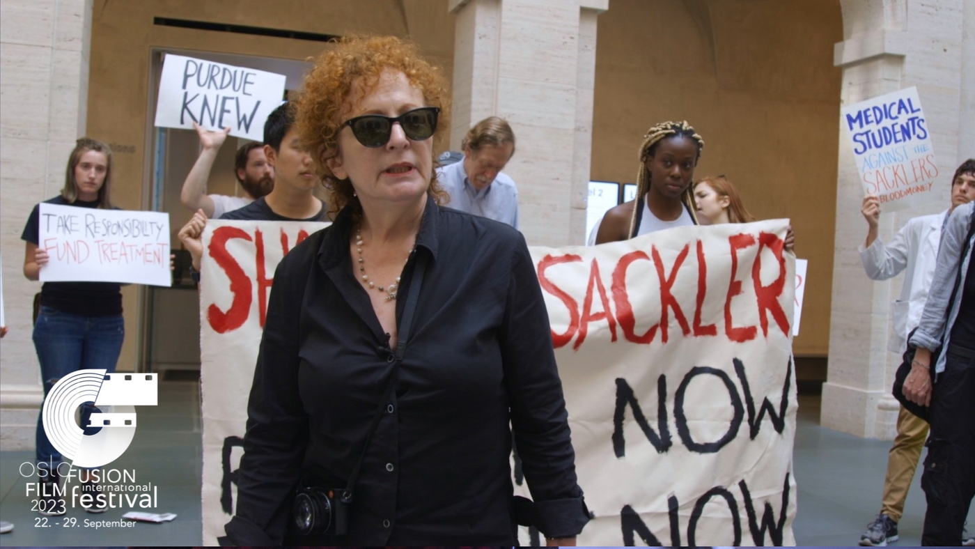 En kvinne står sentrert i bildet med en demonstrasjon i bakgrunn.