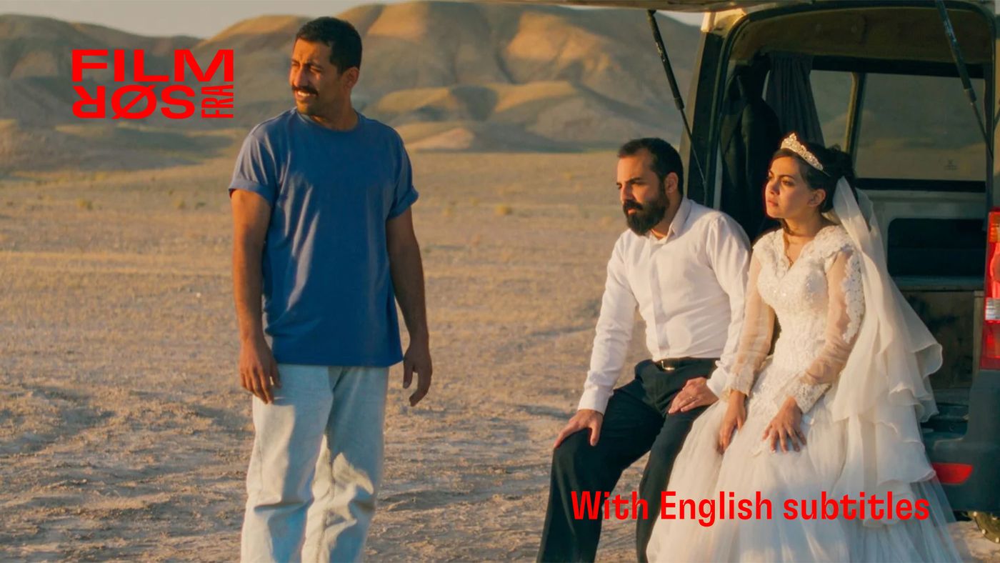 Tre personer ved en varebil i ørkenen. En mann står, en brud og en mann i dress sitter på bagasjekanten.