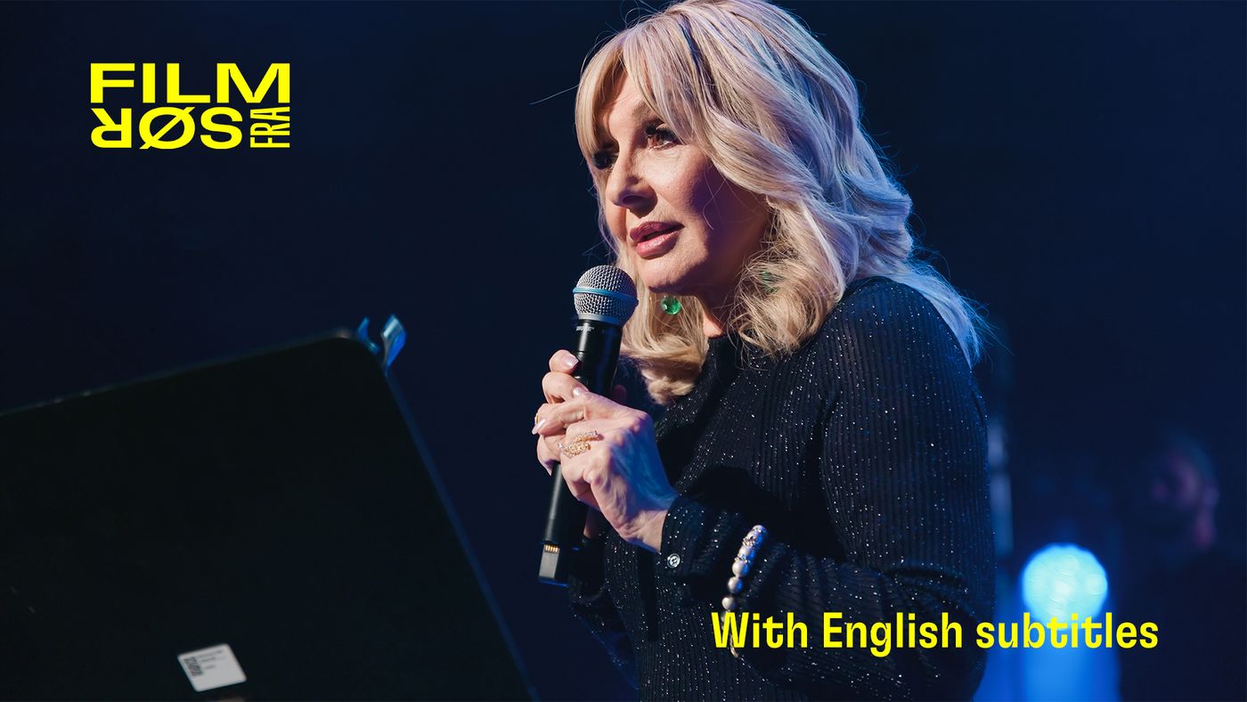 Kvinne med lyst hår og svart kjole står på scene med mikrofon i hånden. Lys og mørk bakgrunn rundt henne.