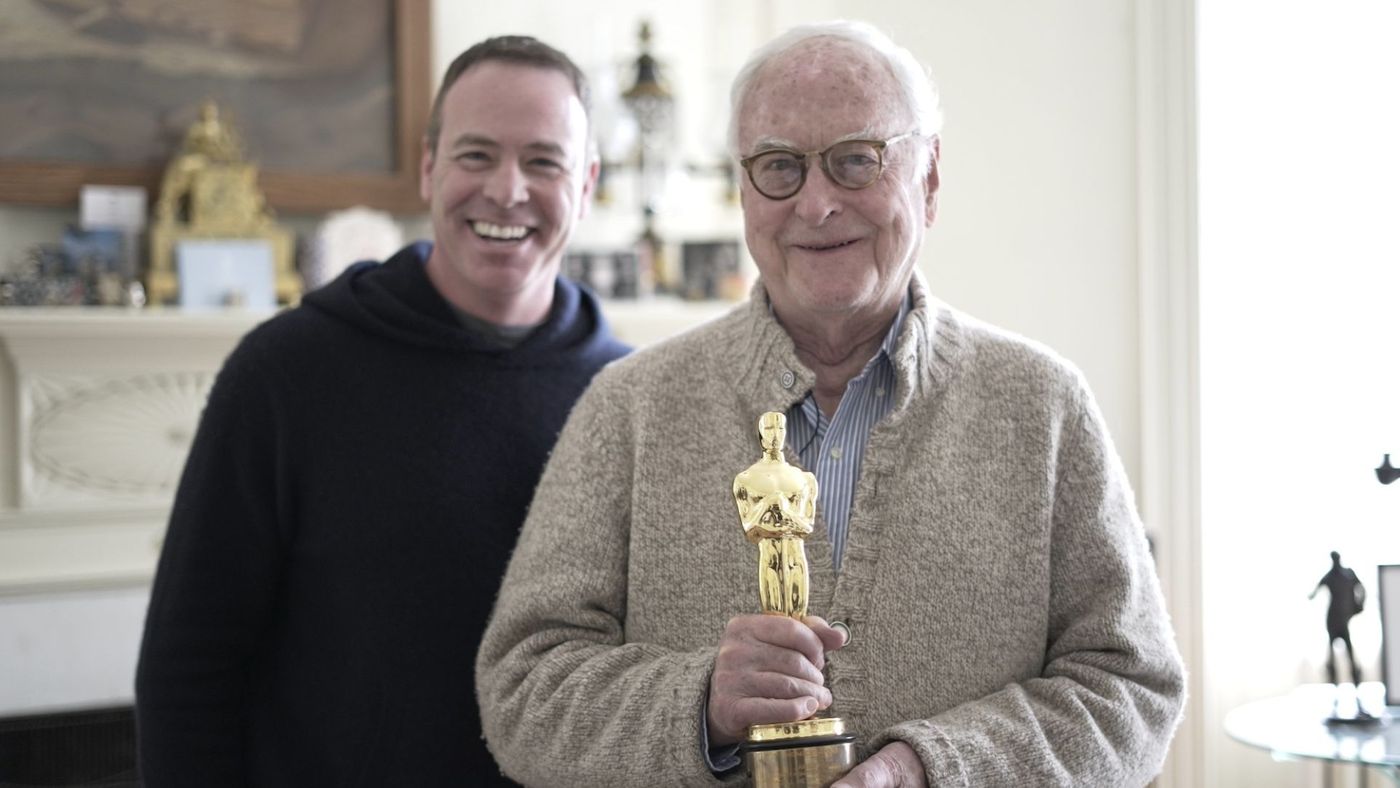 Bildet viser James Ivory som holder hans Oscar statue for manus for Call me by Your Name. Ivory er kledt i en beige ulljakke med blå og hvit stripete skjorte. Han har briller med brun innfatning. I bakgrunnen er en smilende mann kledt i mørk hettegenser. De står foran en peishylle med pyntegjenstander og et maleri.