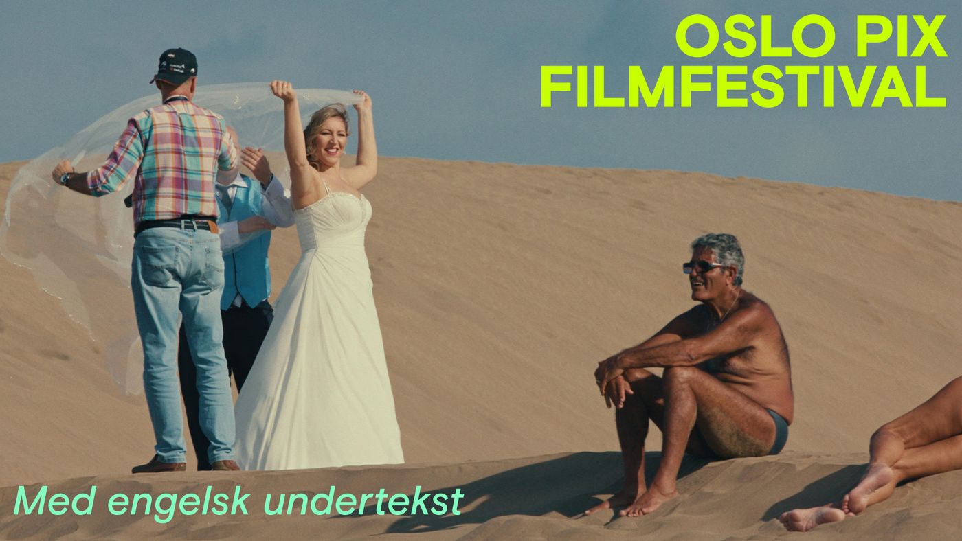 Bildet viser en kvinne i brudekjole som holder sløret over hodet mens hun står i en sanddyne. Ved siden av henne er en mann i en rutete skjorte og jeans som justerer sløret. En annen mann, iført en blå vest og hvit skjorte, står bak bruden. Til høyre i bildet sitter en eldre mann i badebukse på sanden, og en annen person ligger i sanden ved siden av ham. Bakgrunnen er en stor sanddyne under en blå himmel.