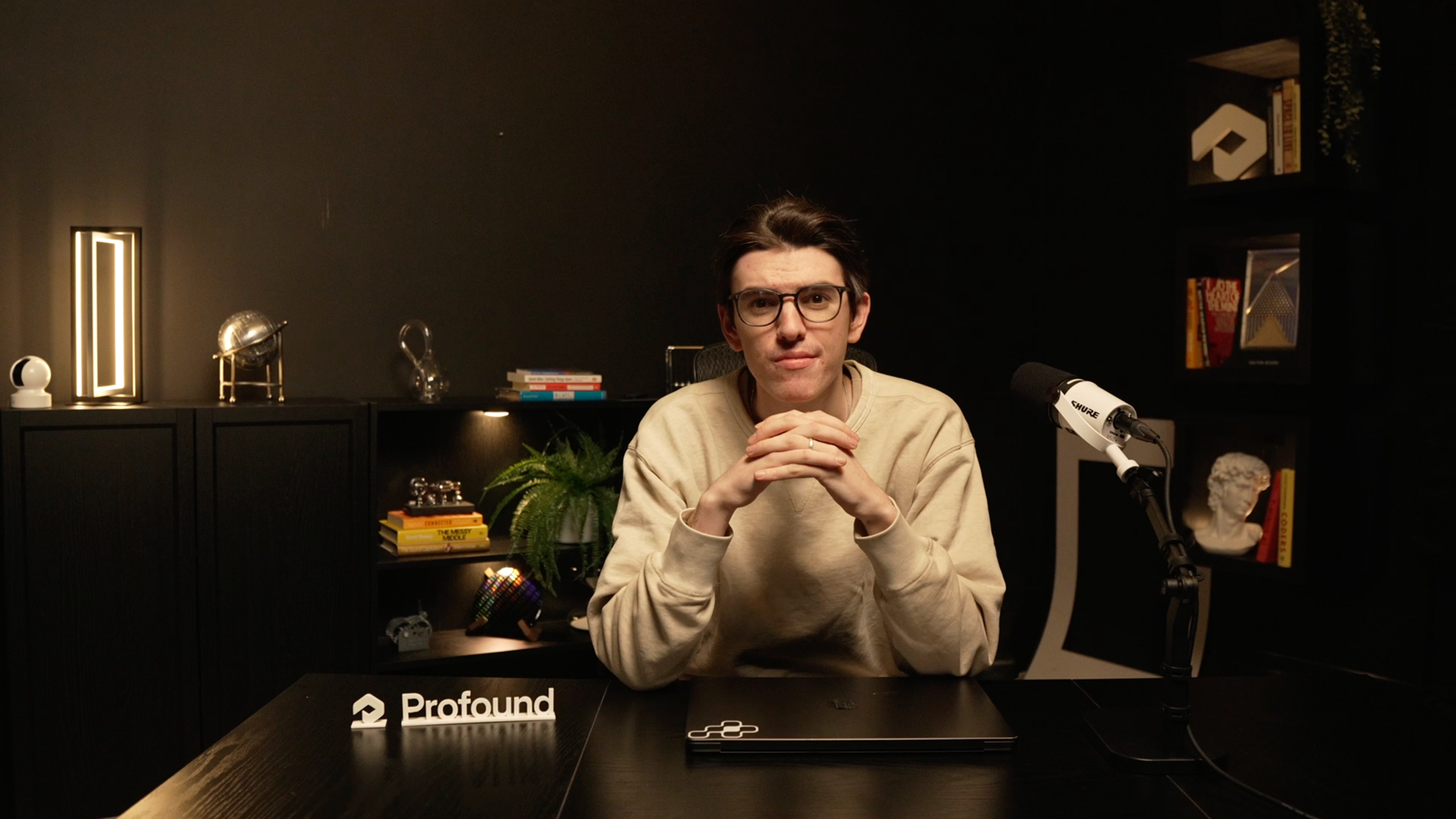 A person sitting at a dark-themed desk with a Profound logo nameplate, a Shure microphone, and a bookshelf in the background, in a professional video recording setup.