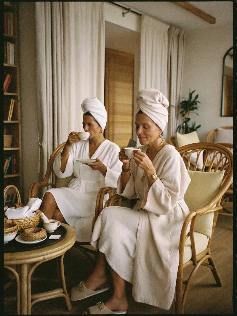 Zwei entspannte Frauen in weißen Bademänteln und mit Handtuch-Turbanen sitzen in gemütlichen Rattansesseln und trinken gemeinsam Tee in einem hellen, einladenden Wellnessbereich.