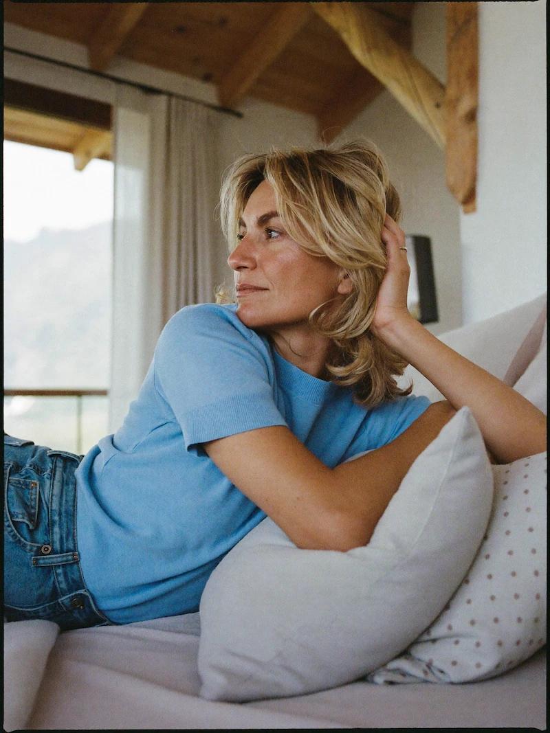 Eine blonde Frau in blauem T-Shirt und Jeans liegt entspannt auf hellen Kissen in einem gemütlichen Zimmer mit rustikaler Holzbalkendecke und blickt nachdenklich aus dem Fenster auf eine Berglandschaft.