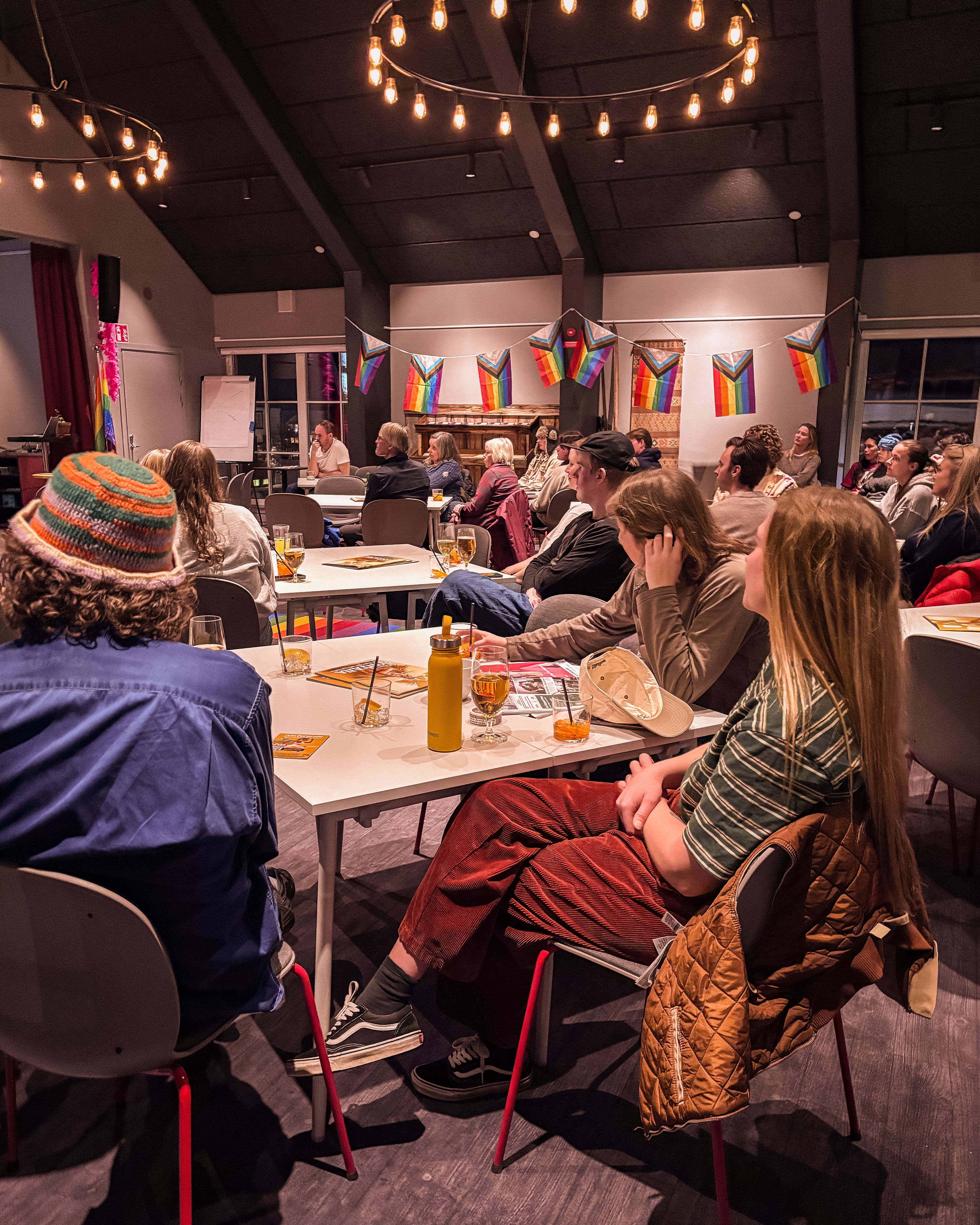 An audience sits at tables in a room decorated with Progress Pride flags and string lights, listening to a speaker.