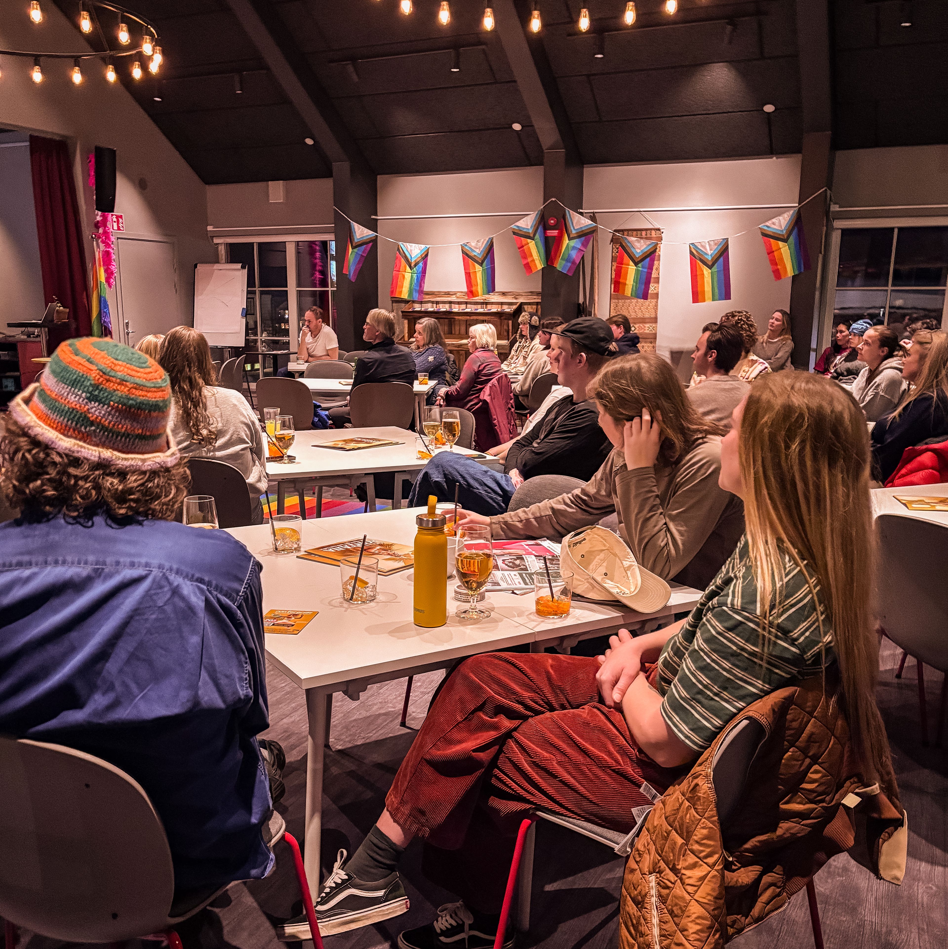 An audience sits at tables in a room decorated with Progress Pride flags and string lights, listening to a speaker.