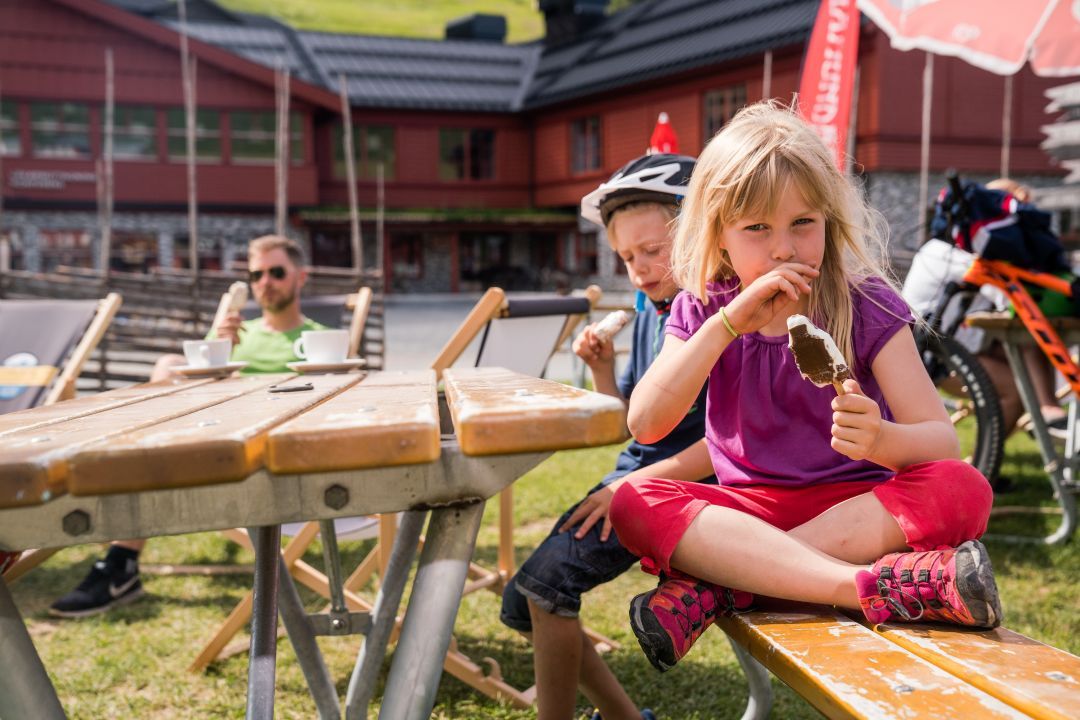Barn som sitter och äter glass på bänk framför en röd byggnad.