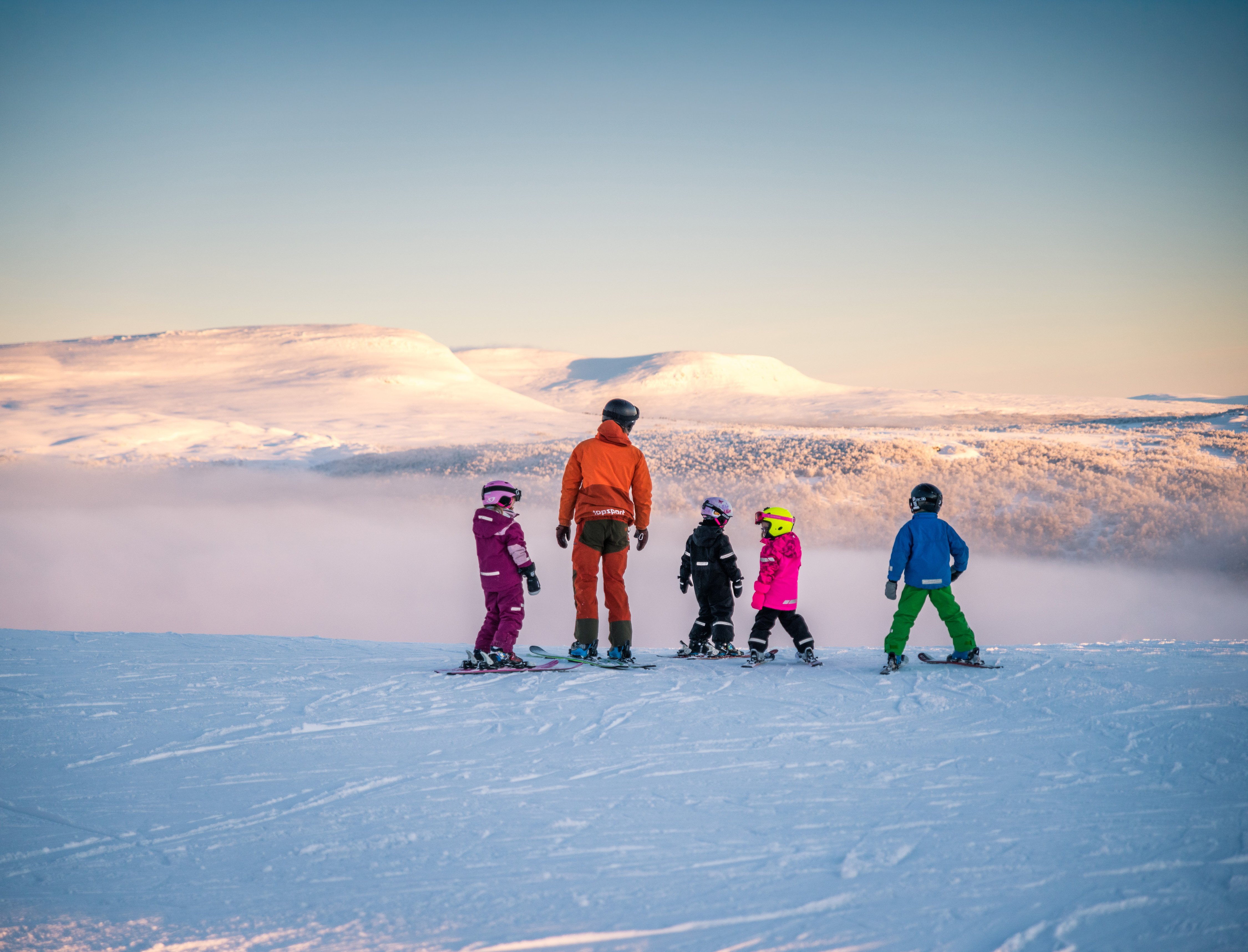 En skidlärare och en grupp barn som står på toppen av en skidbacke.