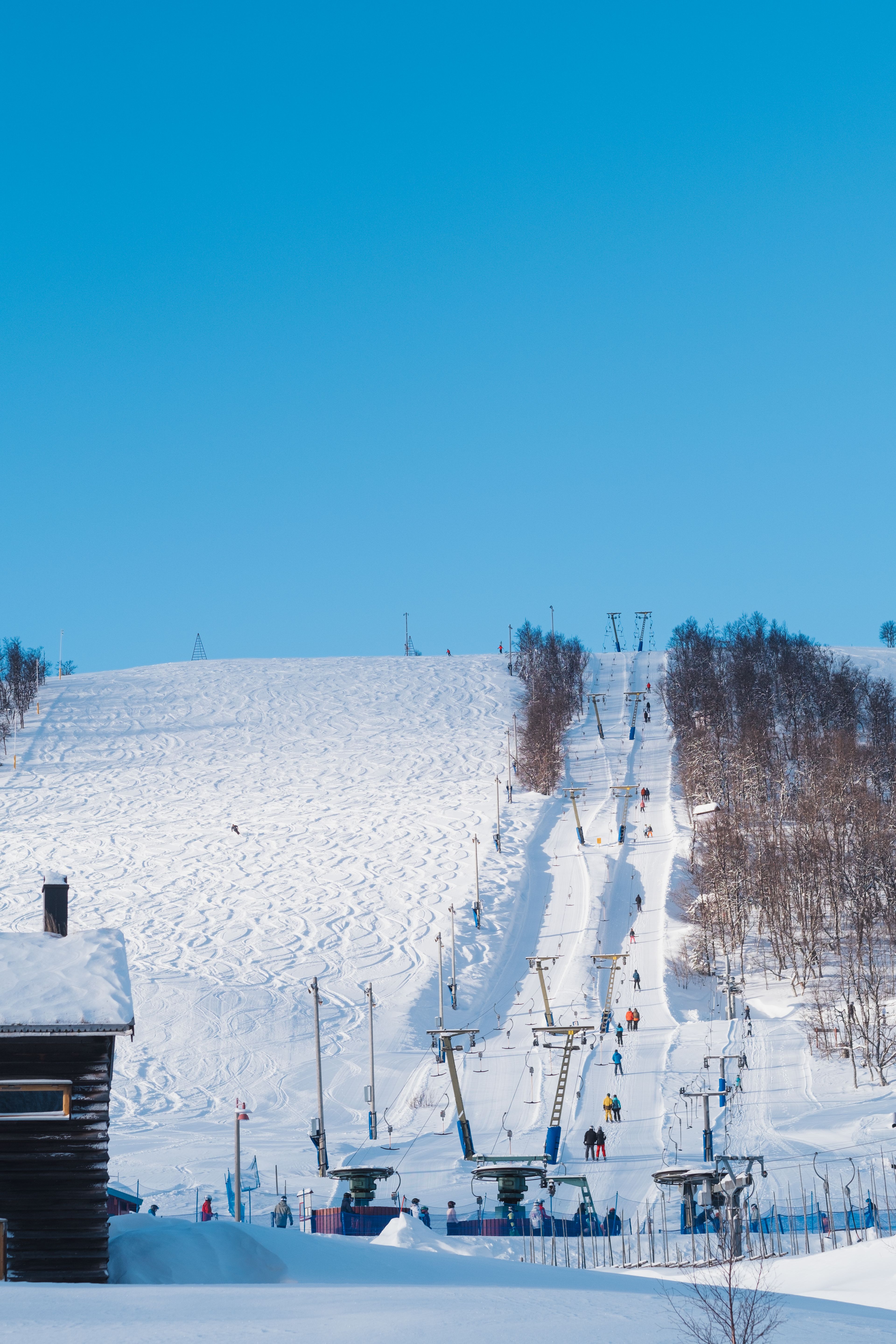 A snowy ski slope with ski lifts, skiers, and ski tracks under a clear blue sky.