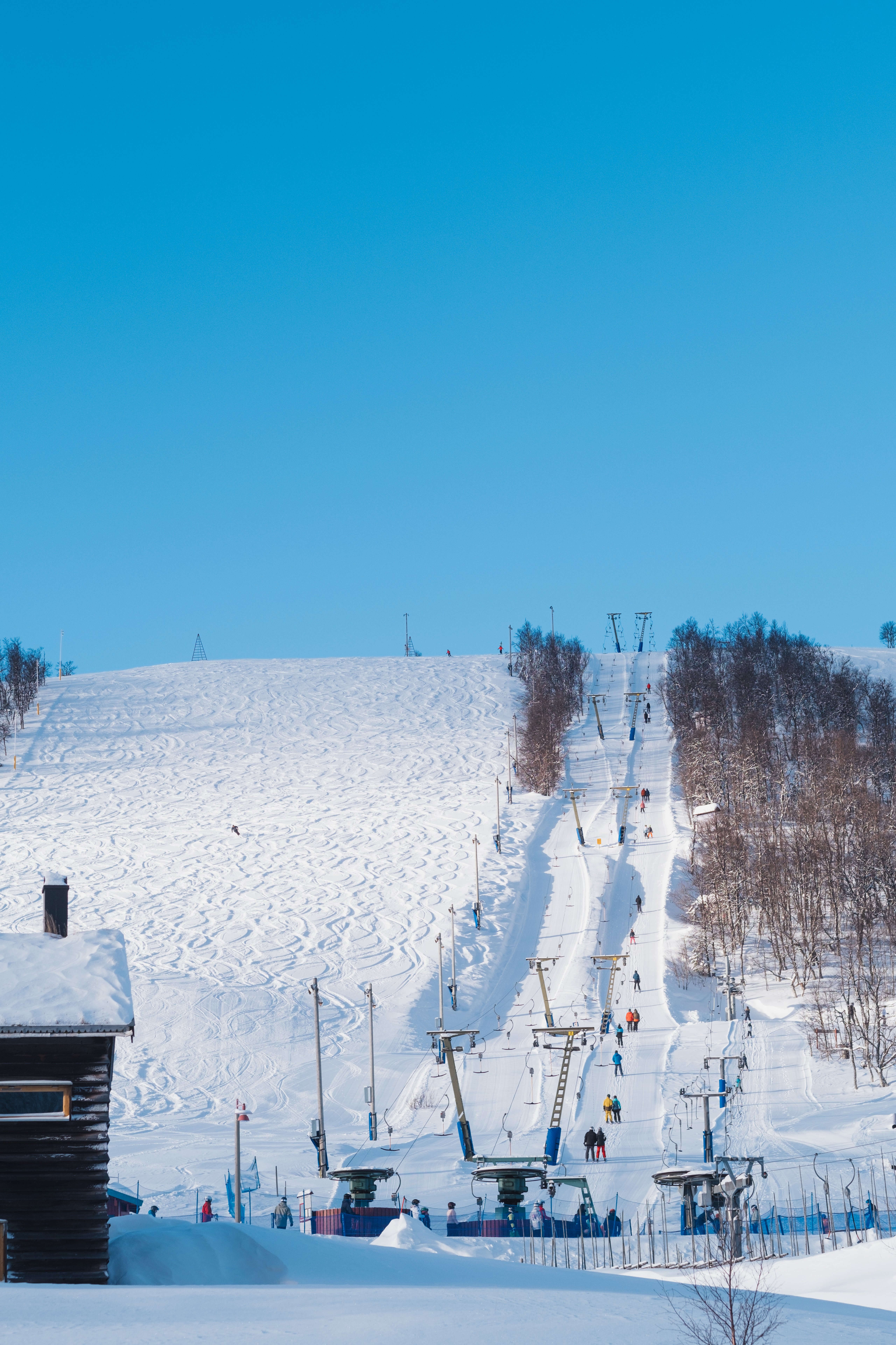 A snowy ski slope with ski lifts, skiers, and ski tracks under a clear blue sky.