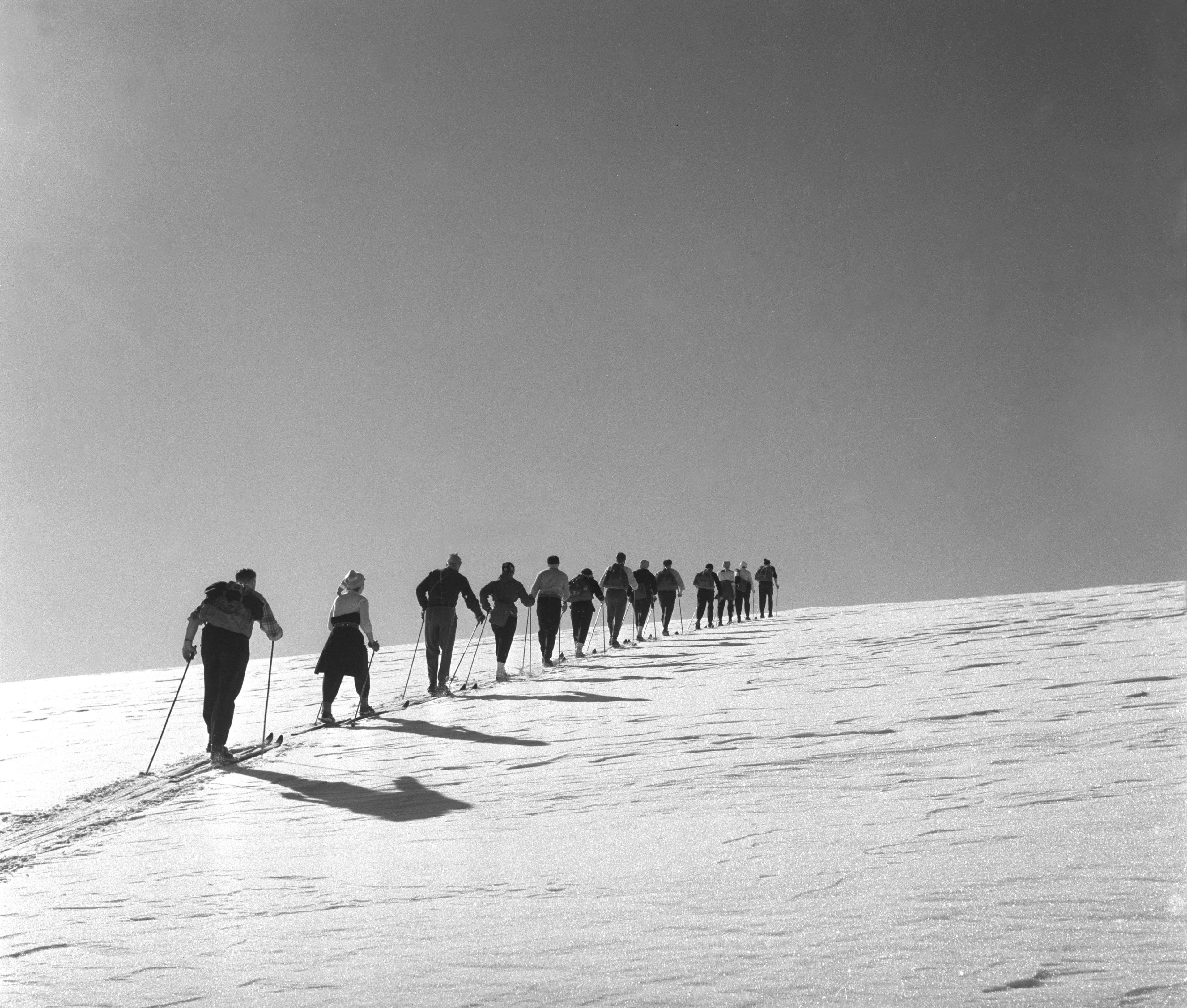 An old photo of a group of people on skis walking up a snow-covered slope.