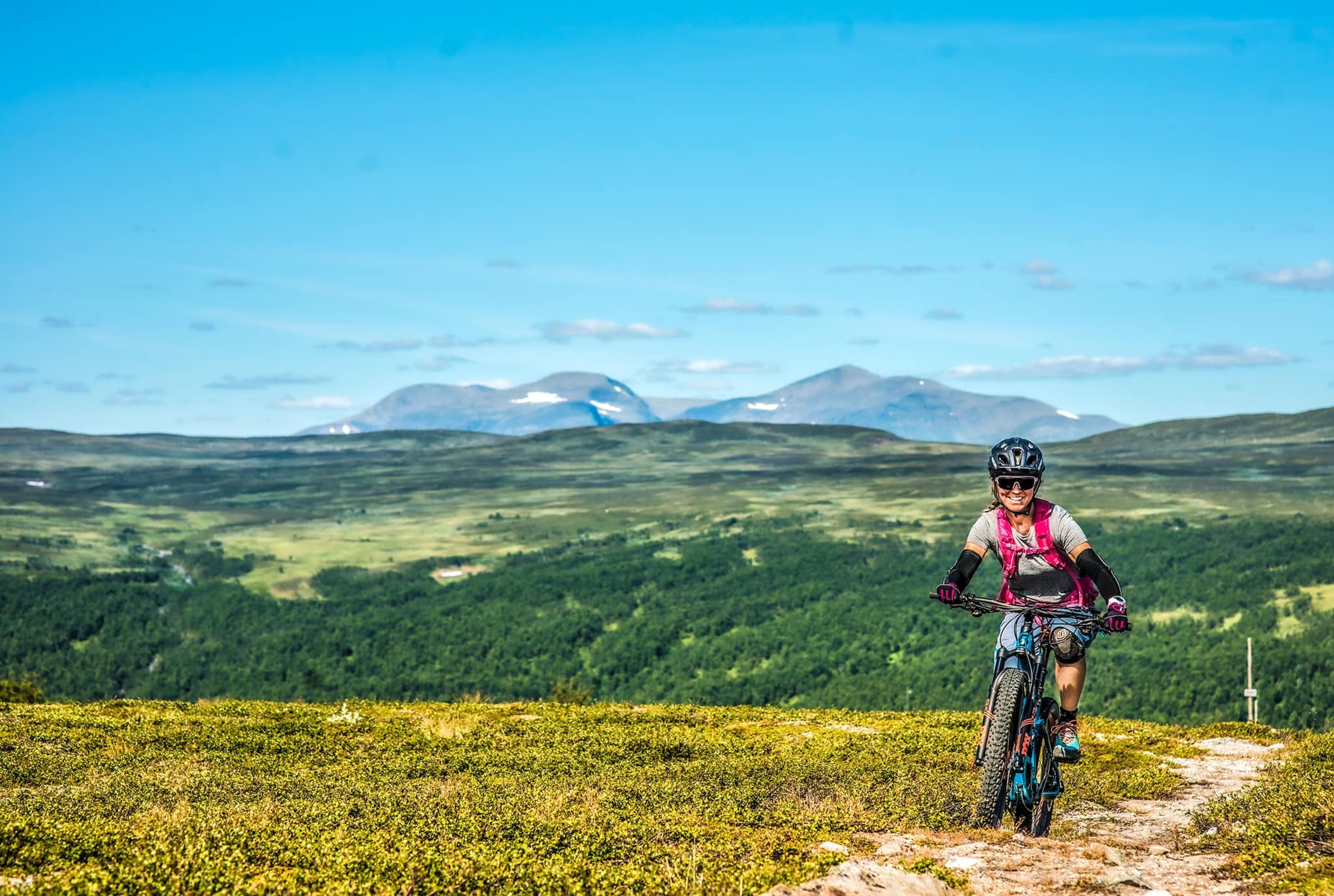 Två personer som står med cyklar på ett fjäll i kvällssjus, med fler fjäll i bakgrunden.