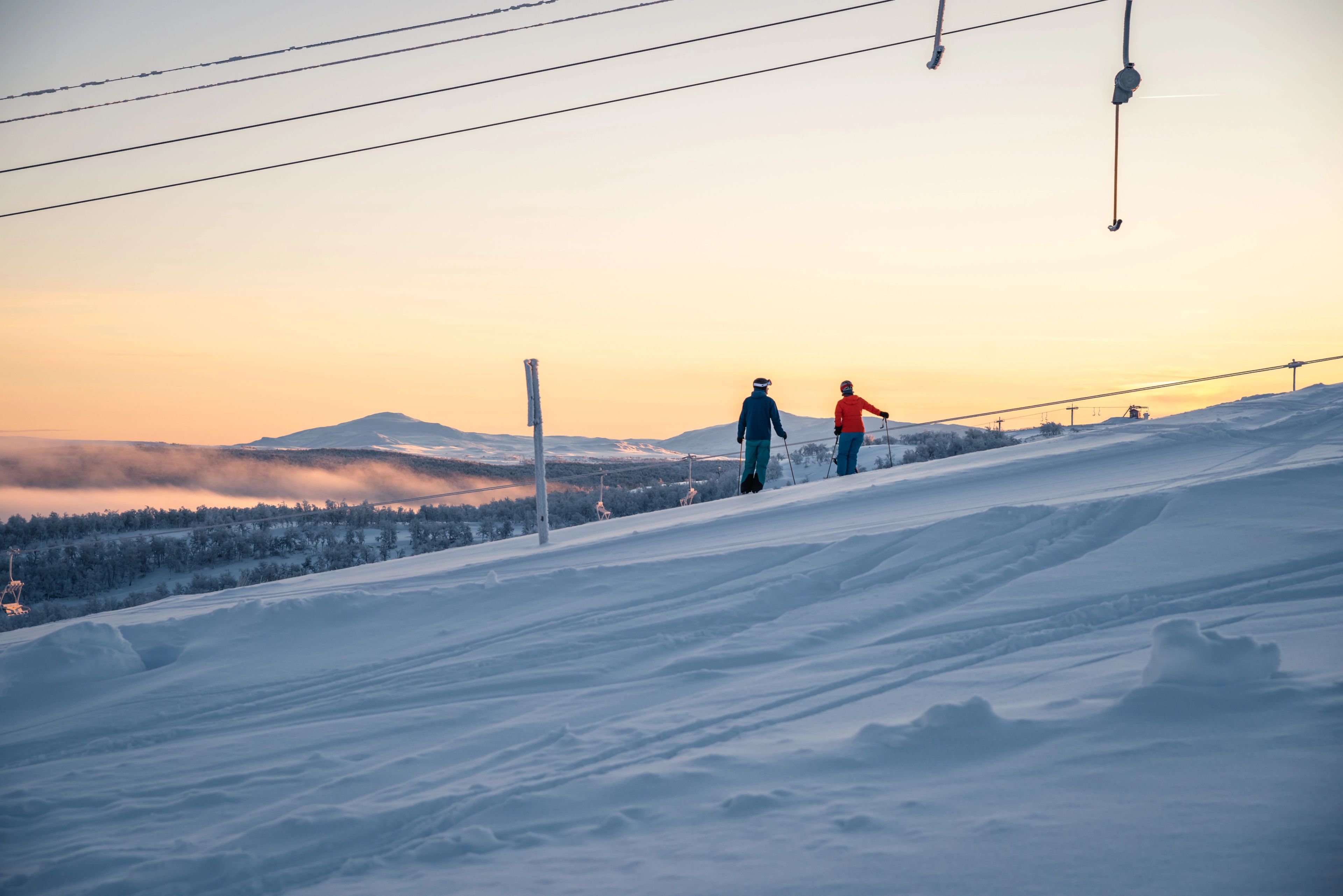 Några personer som står på skidor i solndegången på toppen av en snötäckt sluttning.