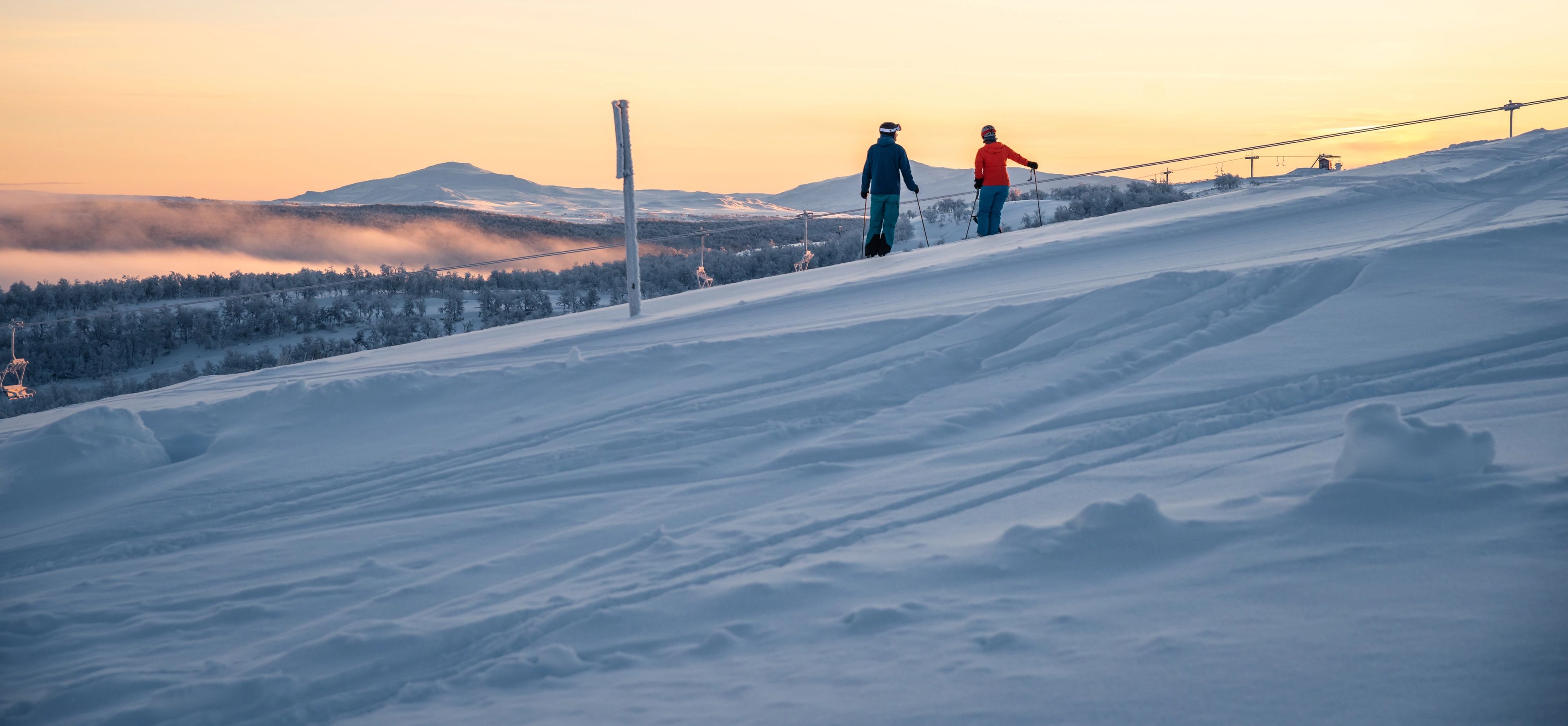 Några personer som står på skidor i solndegången på toppen av en snötäckt sluttning.