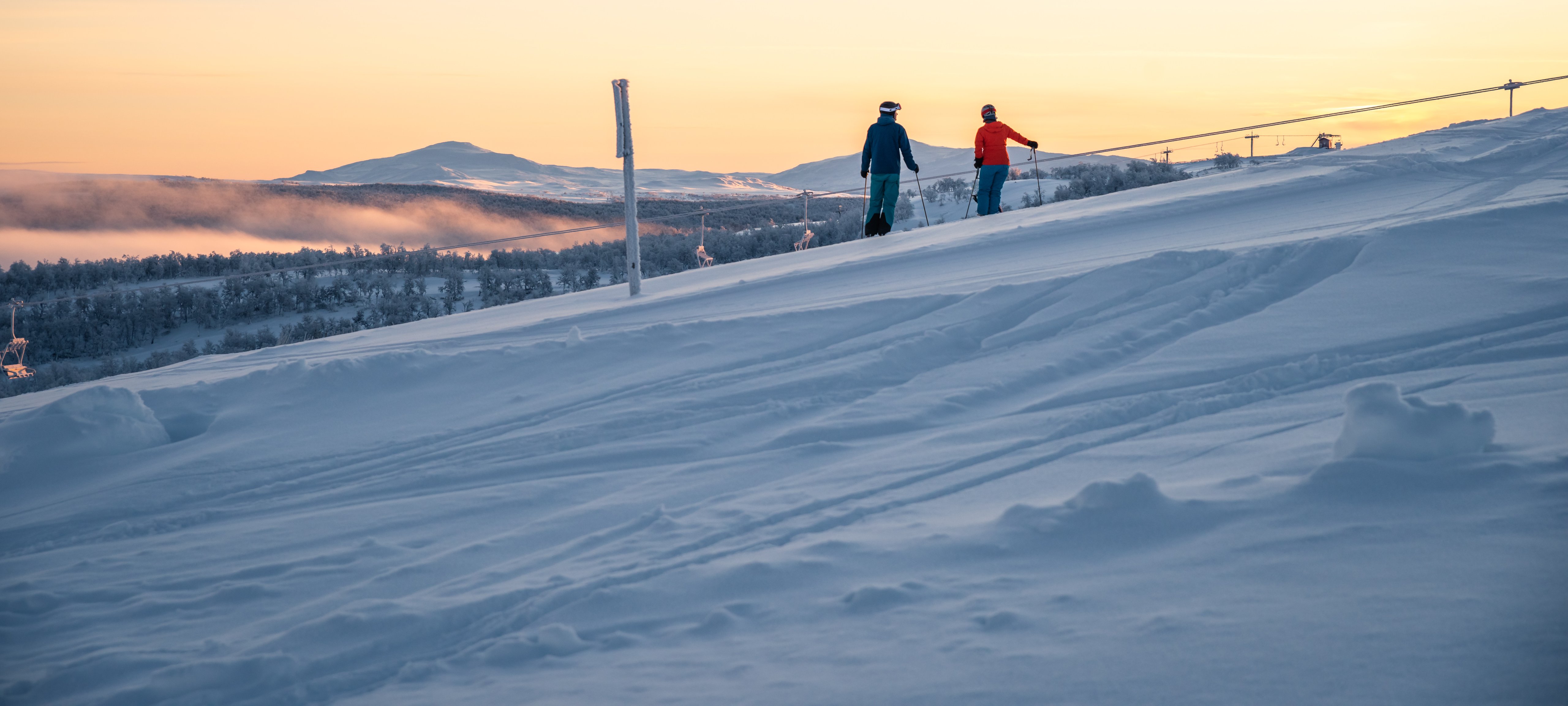Några personer som står på skidor i solndegången på toppen av en snötäckt sluttning.