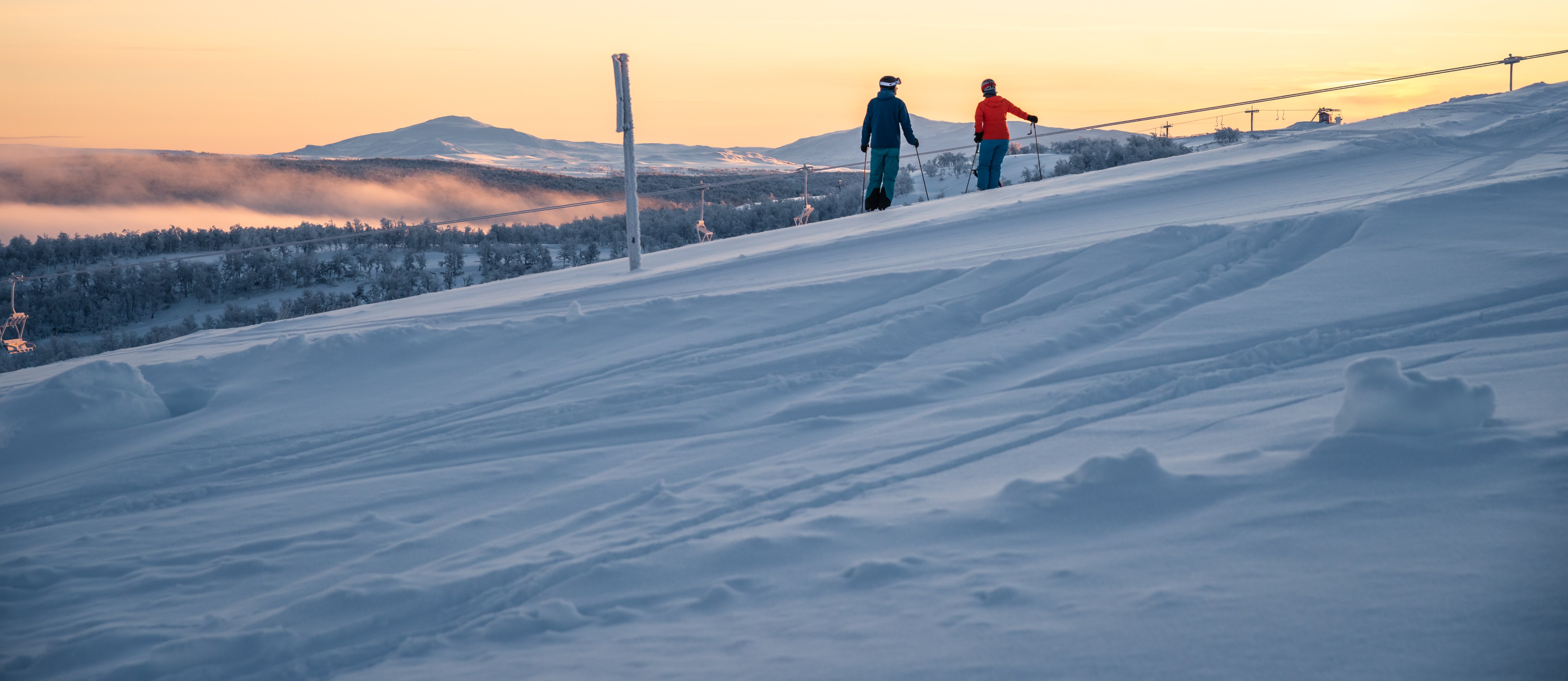 Några personer som står på skidor i solndegången på toppen av en snötäckt sluttning.