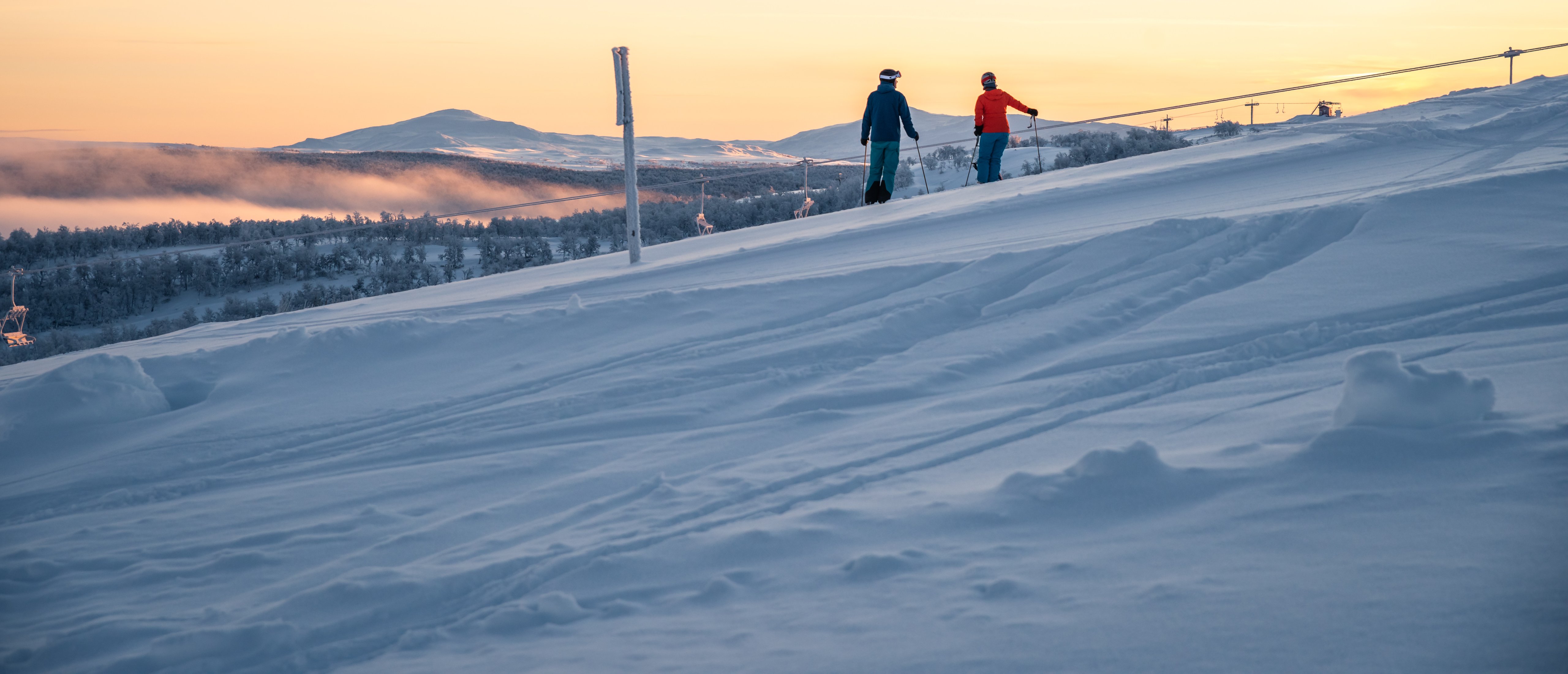 Några personer som står på skidor i solndegången på toppen av en snötäckt sluttning.