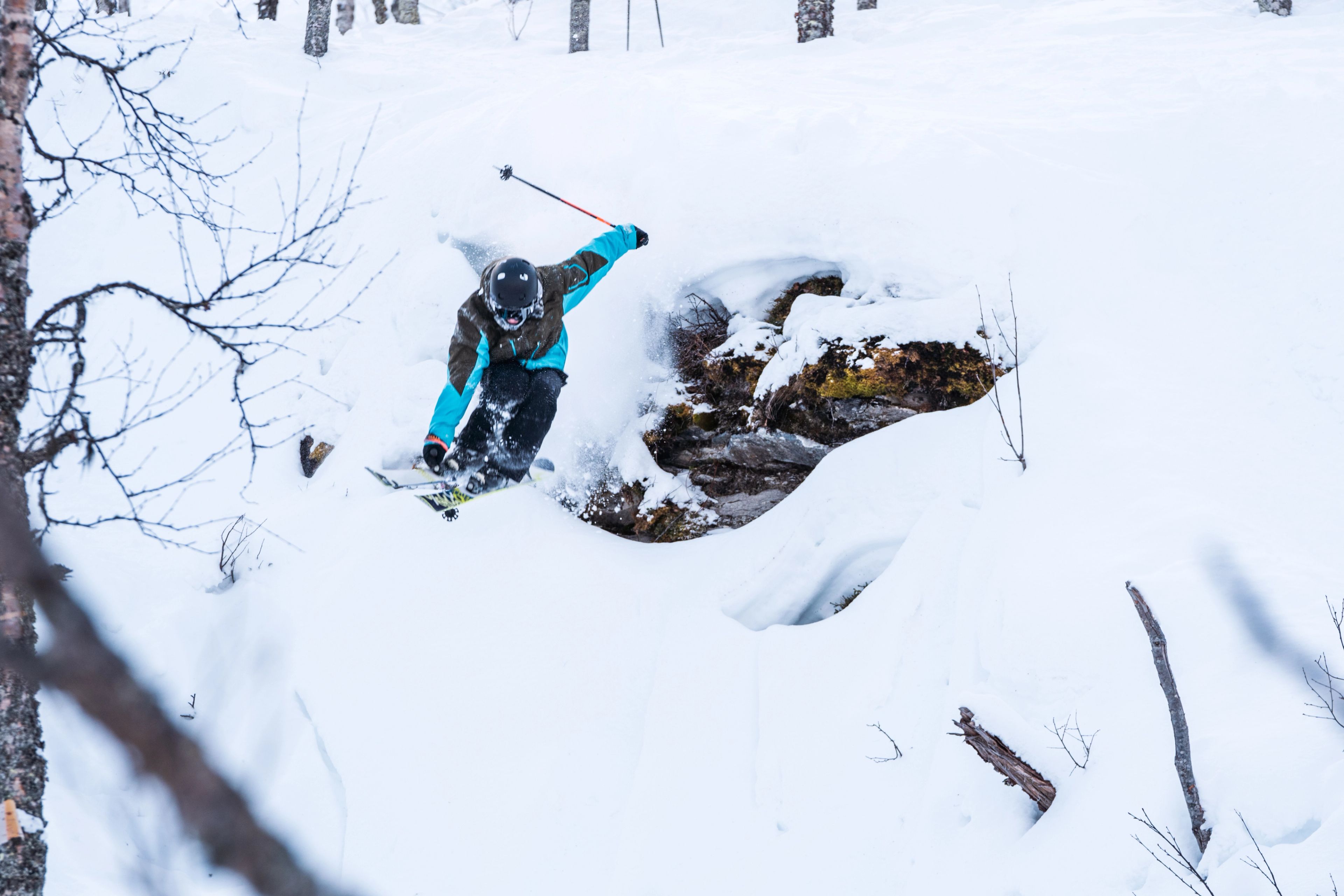 A skier in a brown and teal jacket jumps off a snow-covered rock, kicking up powder in a winter forest.