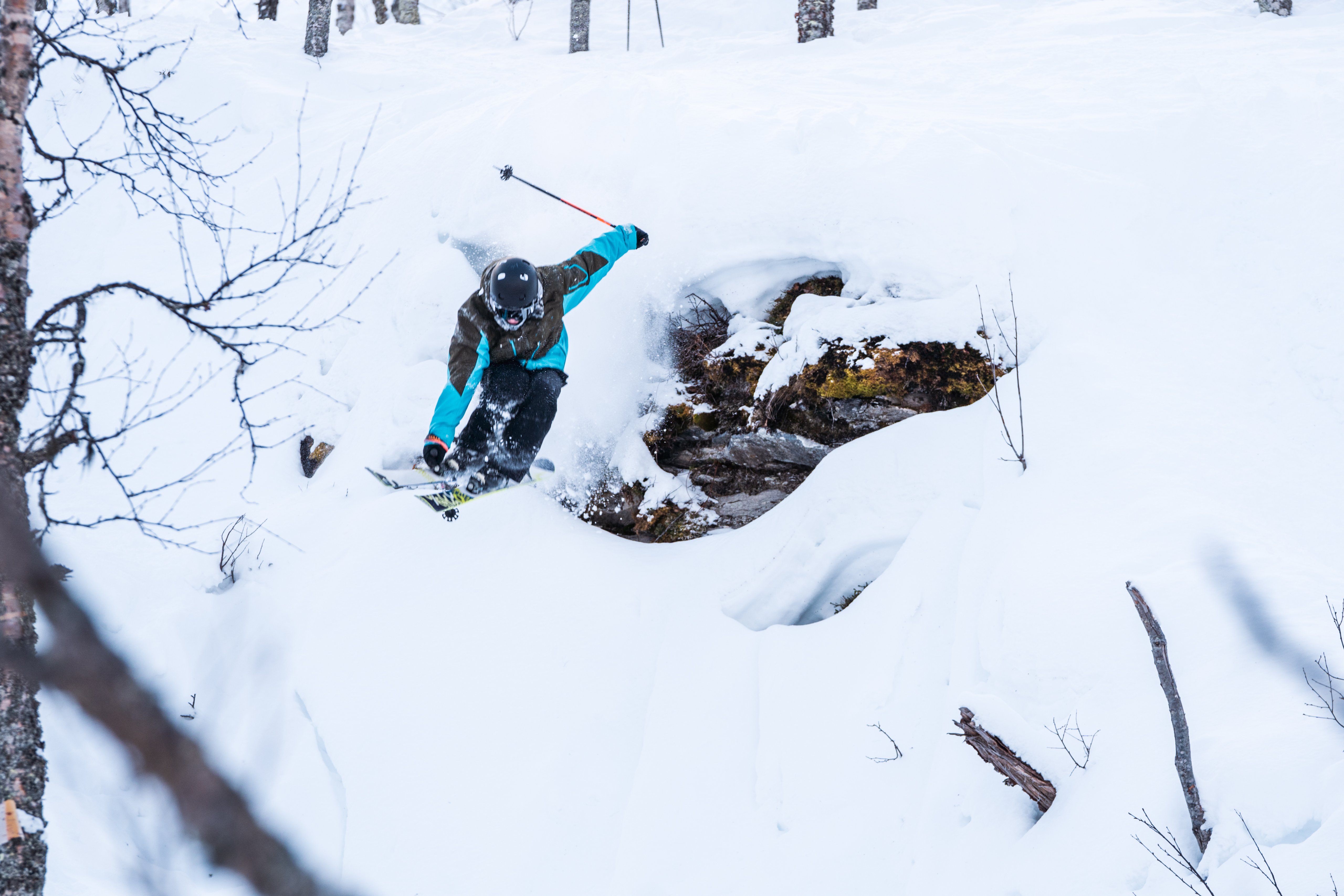 A skier in a brown and teal jacket jumps off a snow-covered rock, kicking up powder in a winter forest.