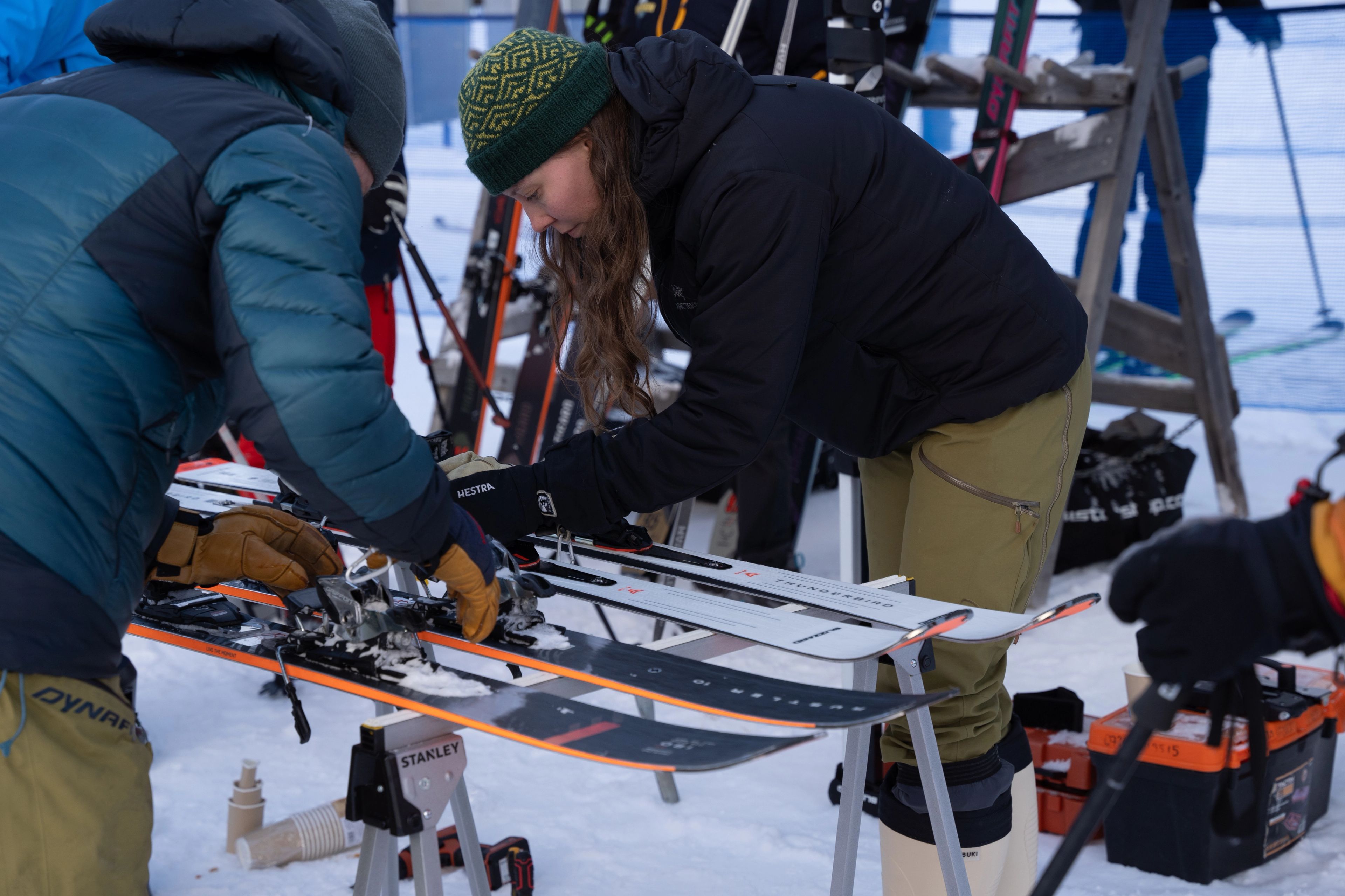 A woman in a black jacket and a man in a blue jacket adjusting a pair of skis.