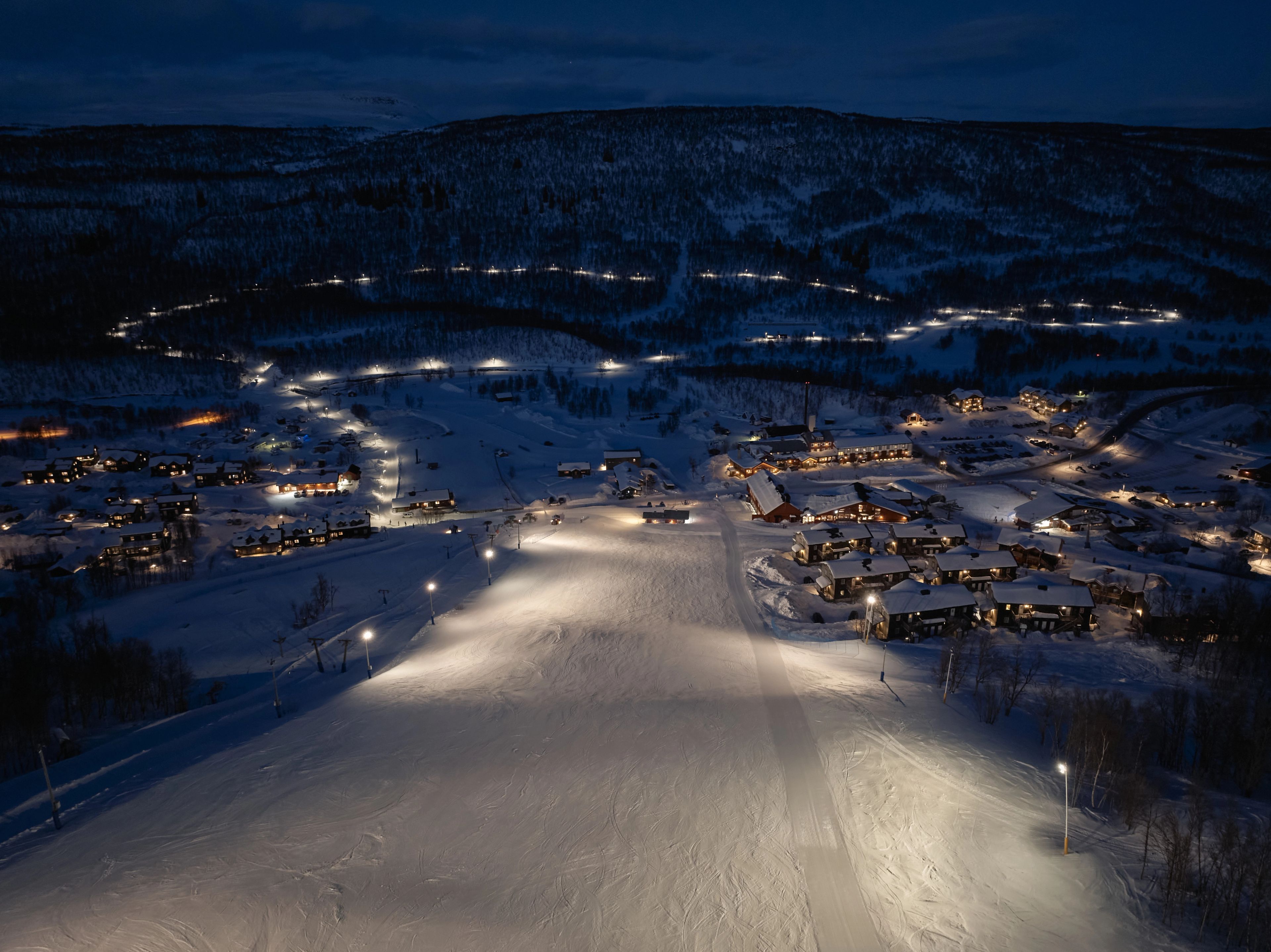 A skier descends an illuminated slope at a snowy ski resort with multiple lit buildings.