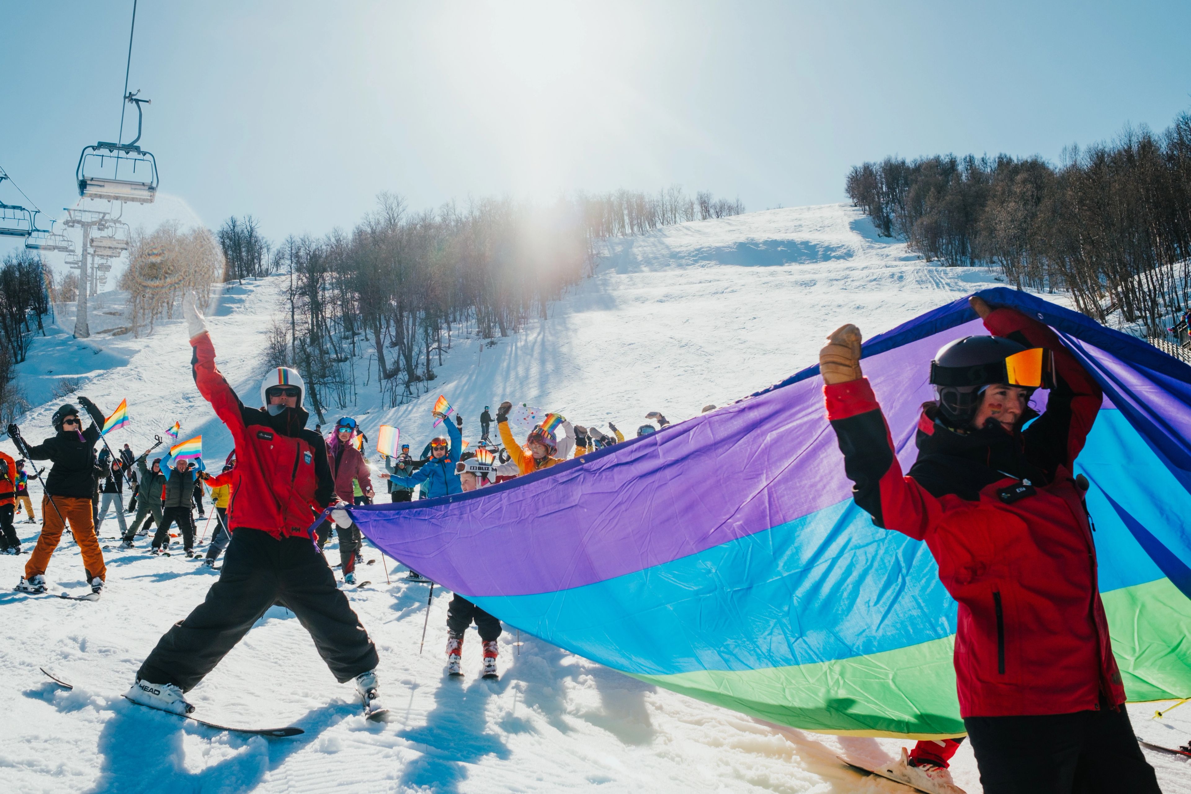 A group of skiers on a sunny mountain holds a large, colorful flag and smaller pride flags.
