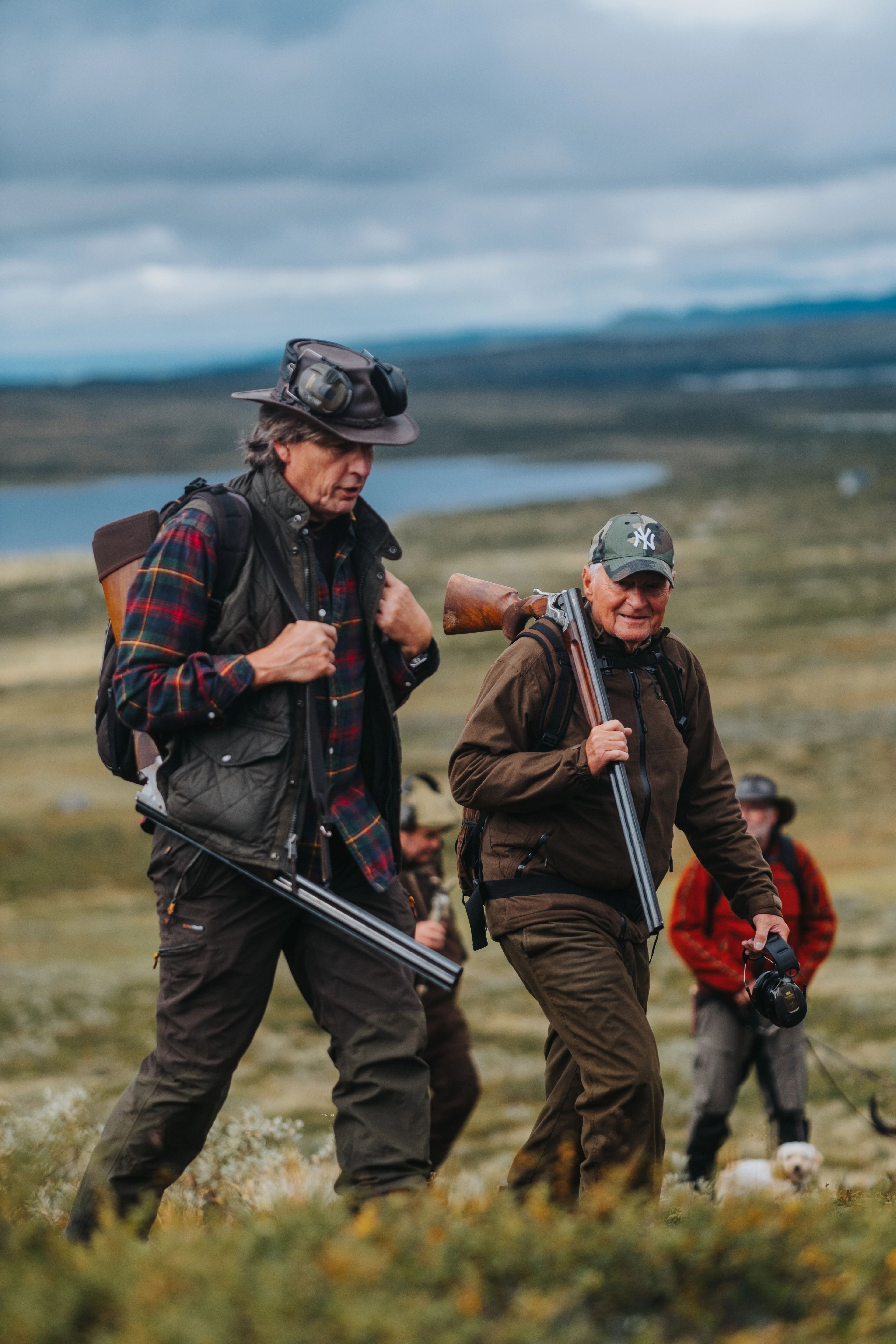 Två män som deltar i en ripjakt går upp för en fjällsluttning med en sjö i bakgrunden och ytterligare en manlig jaktdeltagare.
