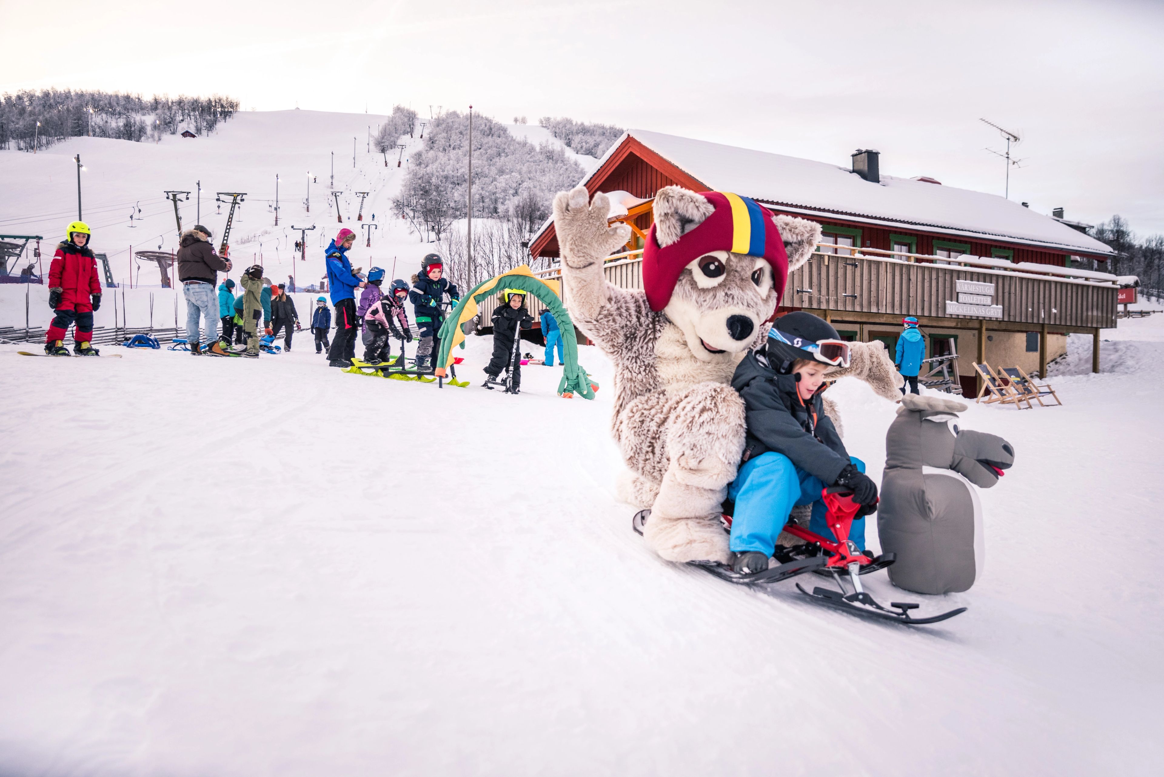 En barn som åker pulka tillsammans med en maskot nedför en snötäckt sluttning. 
