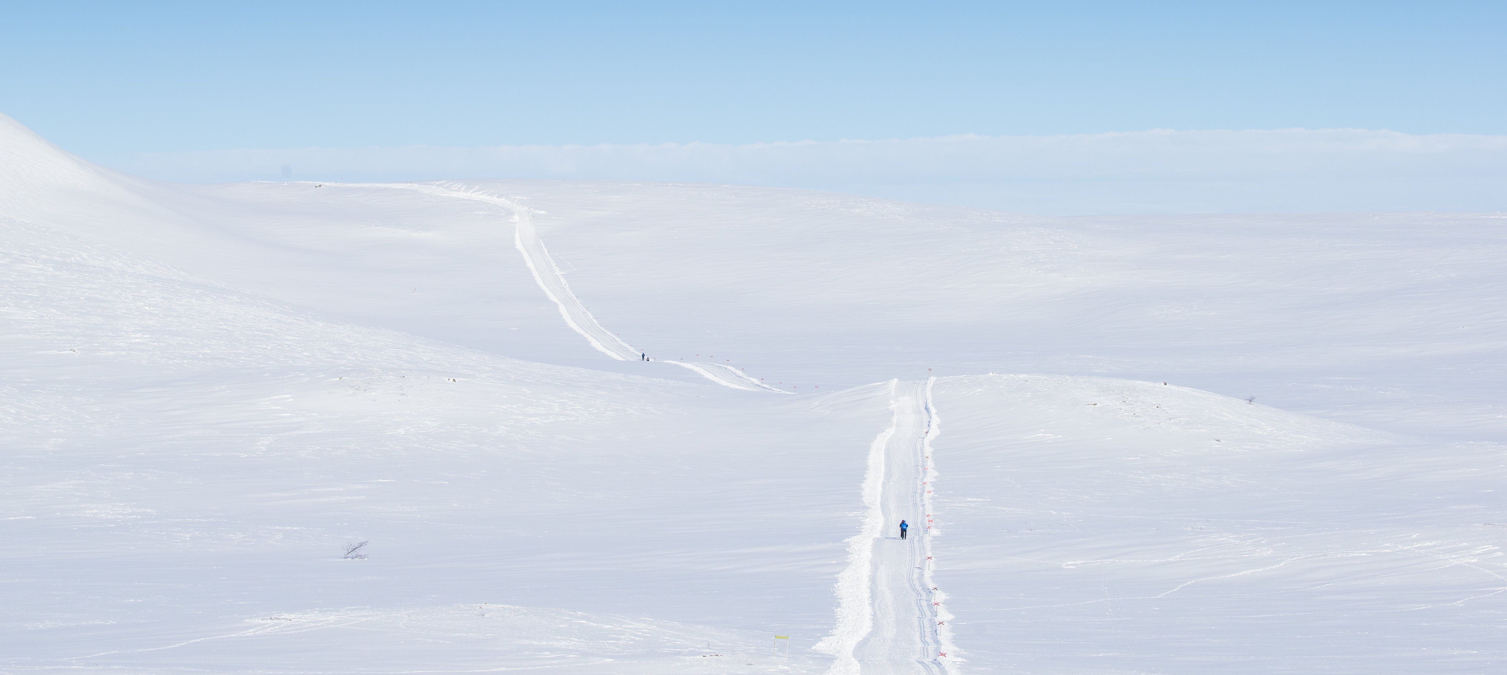 Person ski on a snowy path through a vast white landscape under a blue sky.