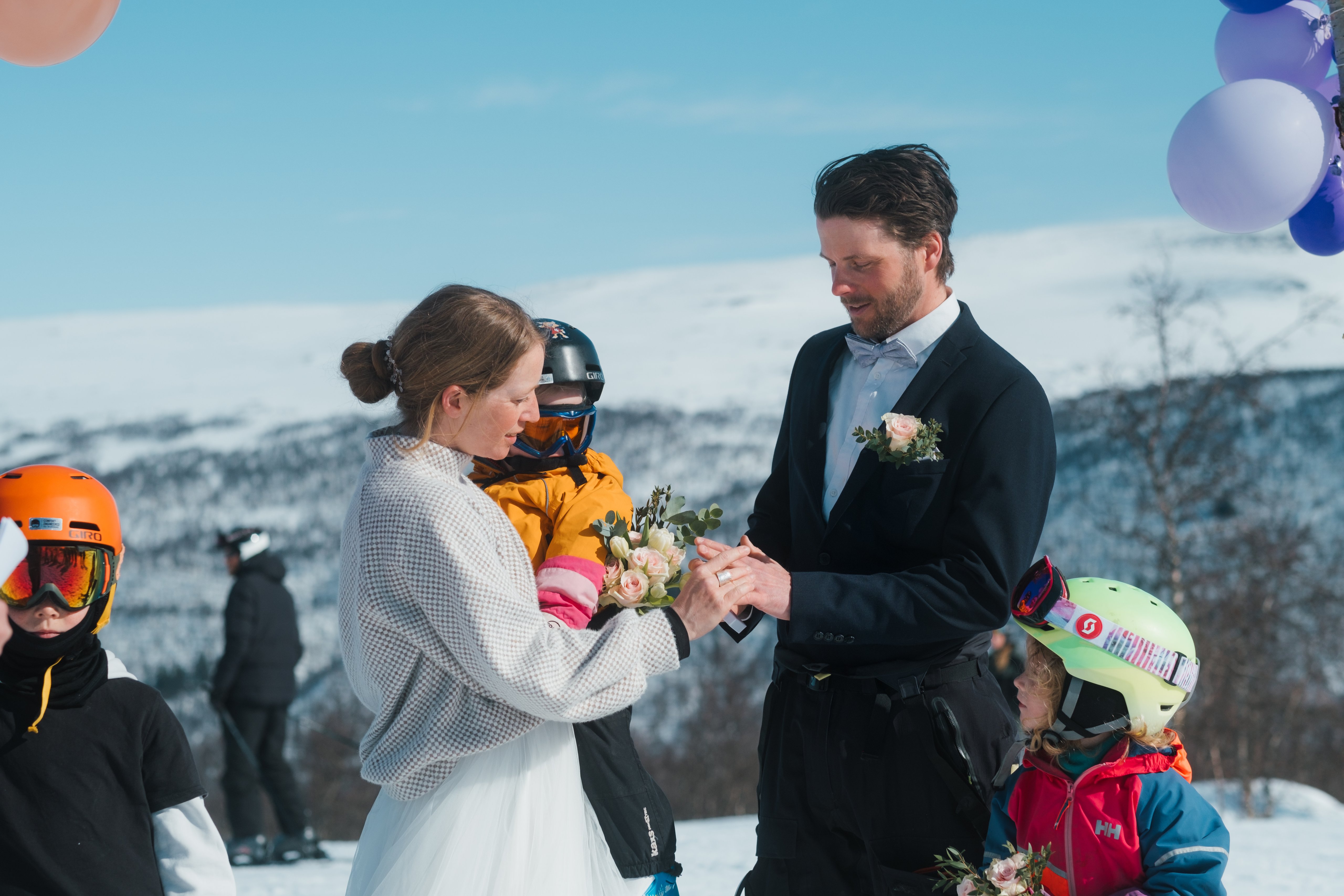 Bride and groom exchange rings at a snowy mountain wedding, with children in ski gear nearby.