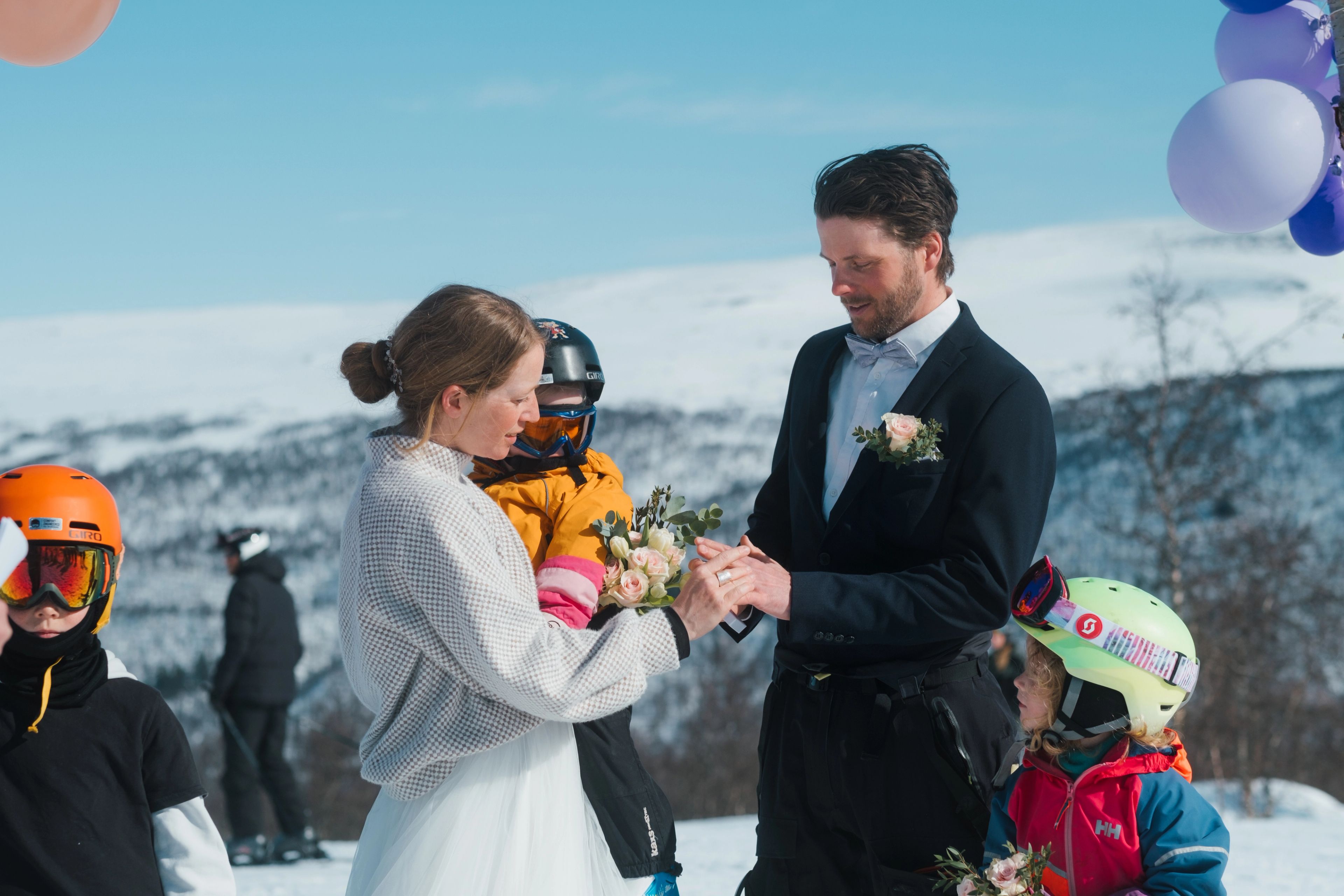 Bride and groom exchange rings at a snowy mountain wedding, with children in ski gear nearby.