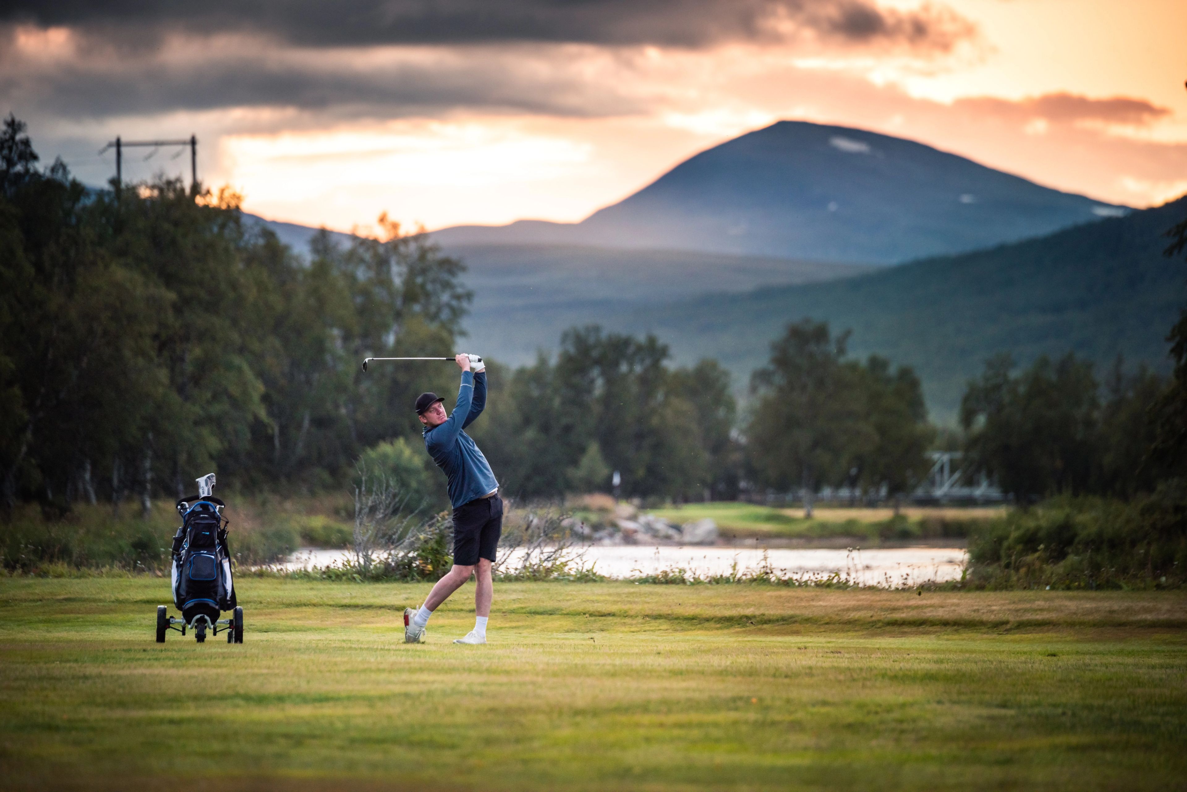 A man swings a golf club on a course with a lake, trees, and mountains under a sunset sky.
