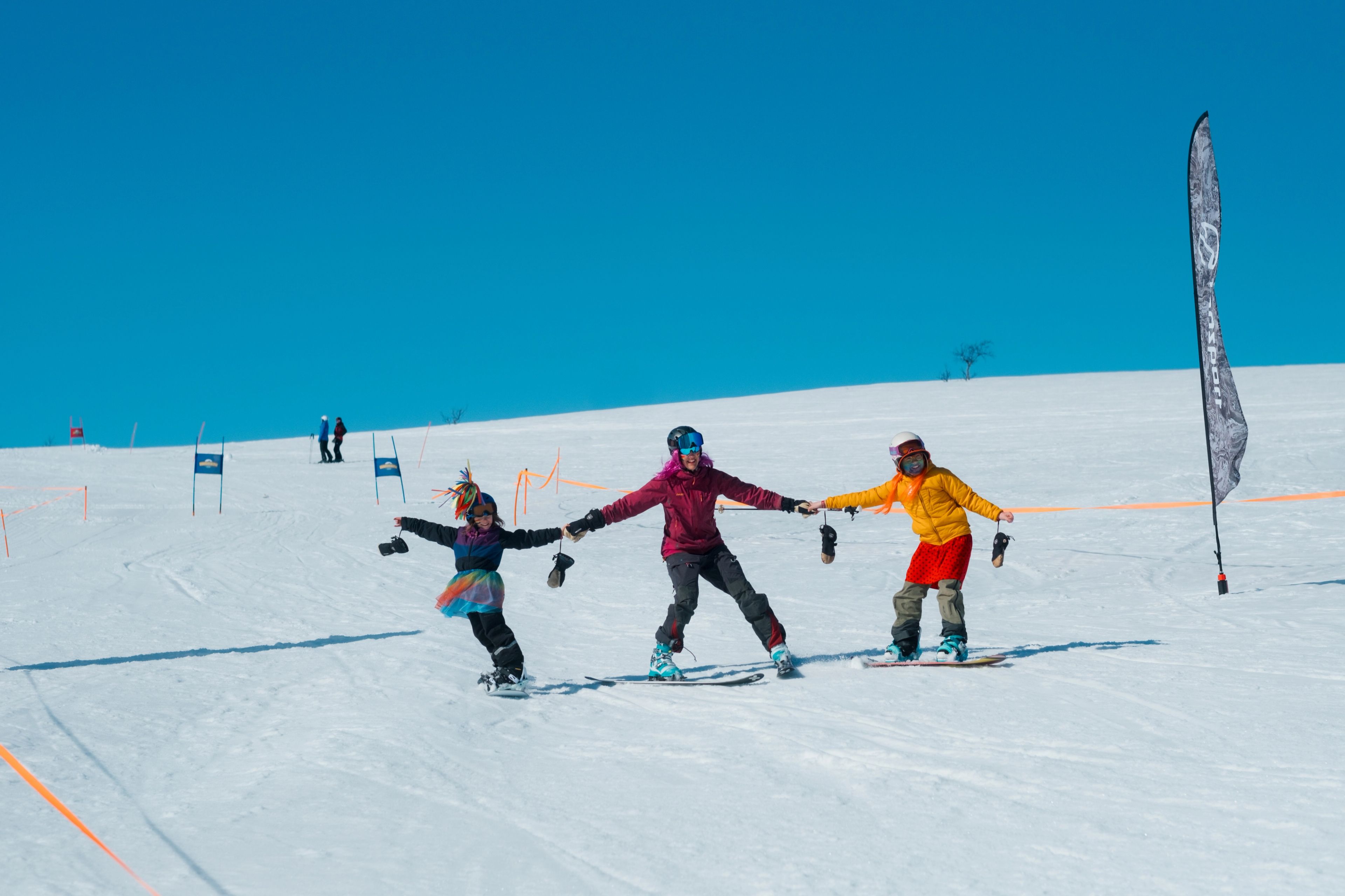 Three people, two on snowboards and one on skis, hold hands while descending a snowy slope under a clear blue sky.