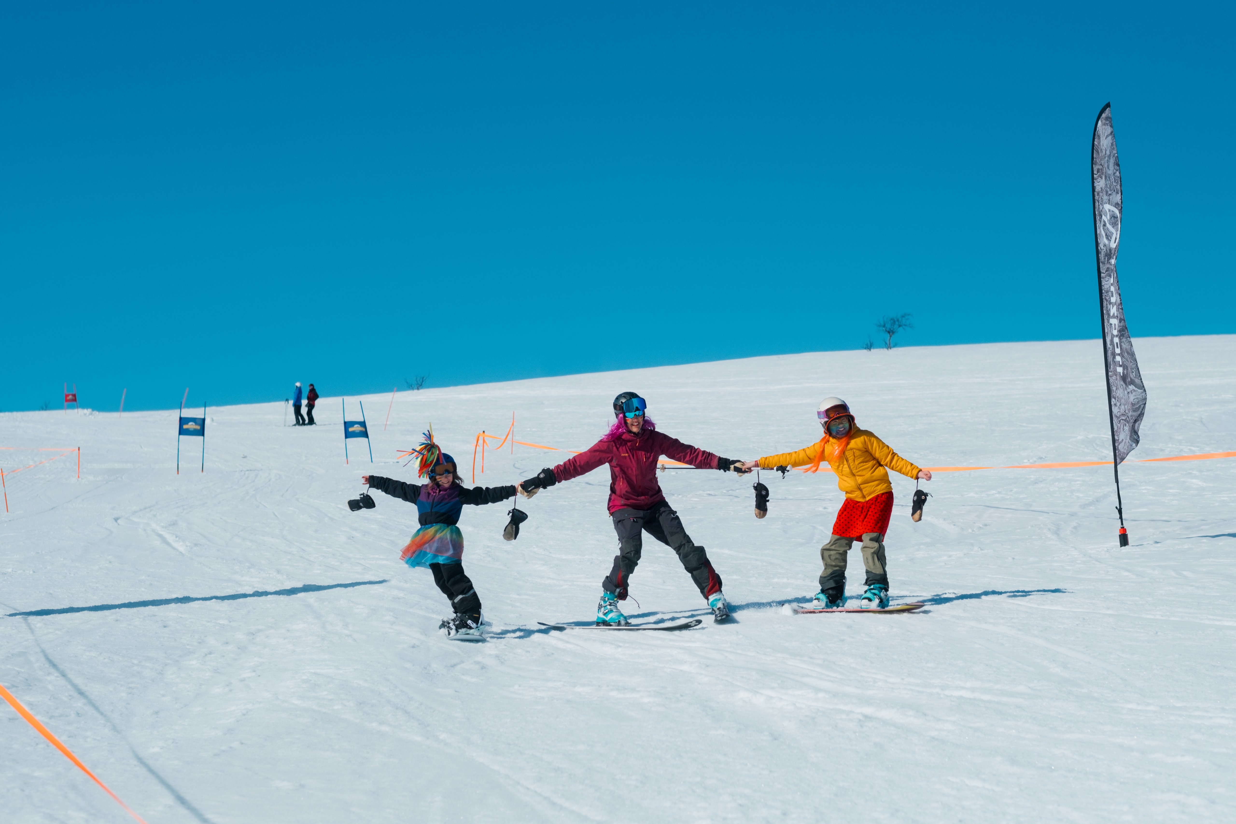 Three people, two on snowboards and one on skis, hold hands while descending a snowy slope under a clear blue sky.