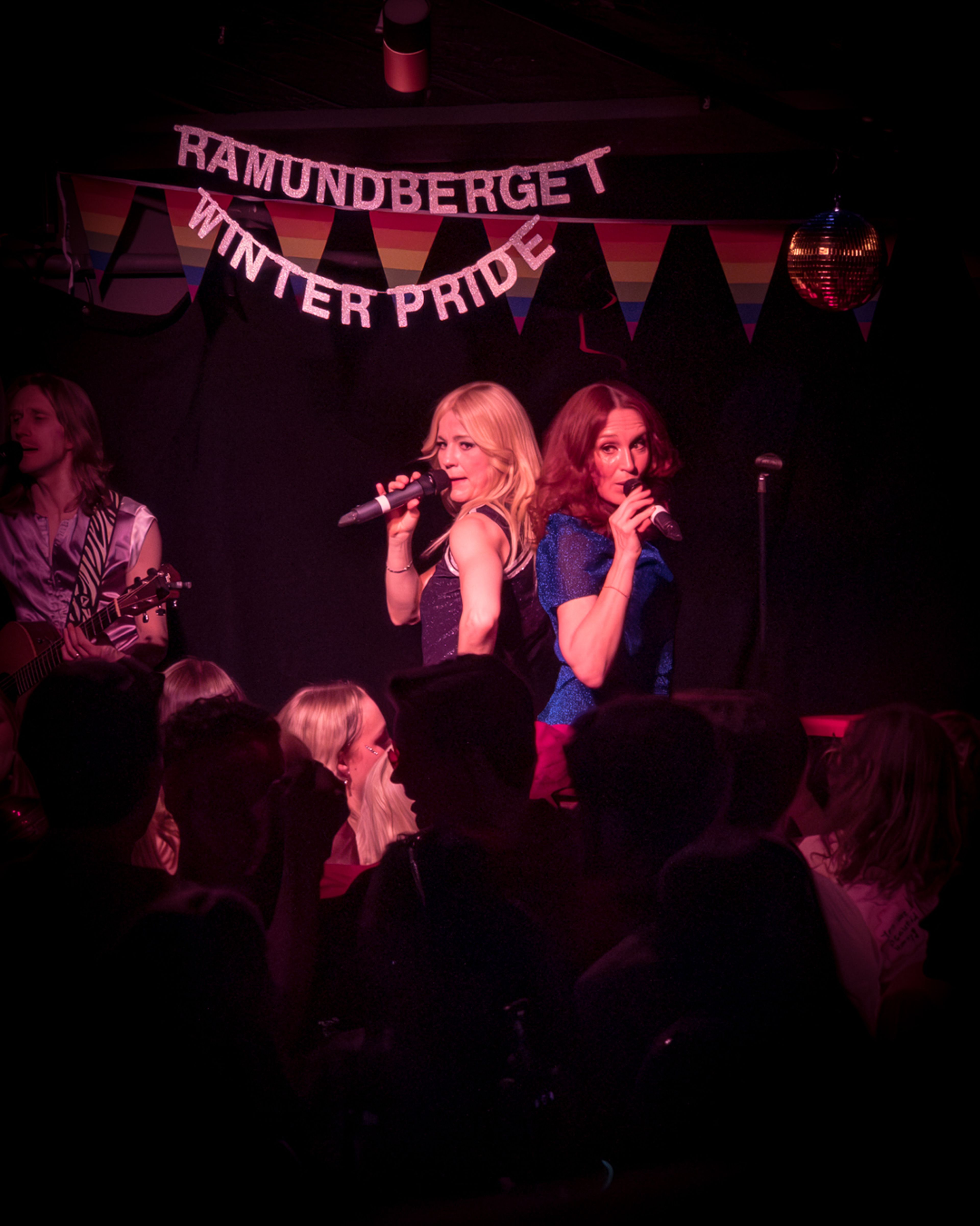 Two women sing into microphones on a dark stage under pink lights, with a guitarist behind them and an audience in silhouette. A banner reads "RAMUNDBERGET WINTER PRIDE" above a rainbow flag bunting.