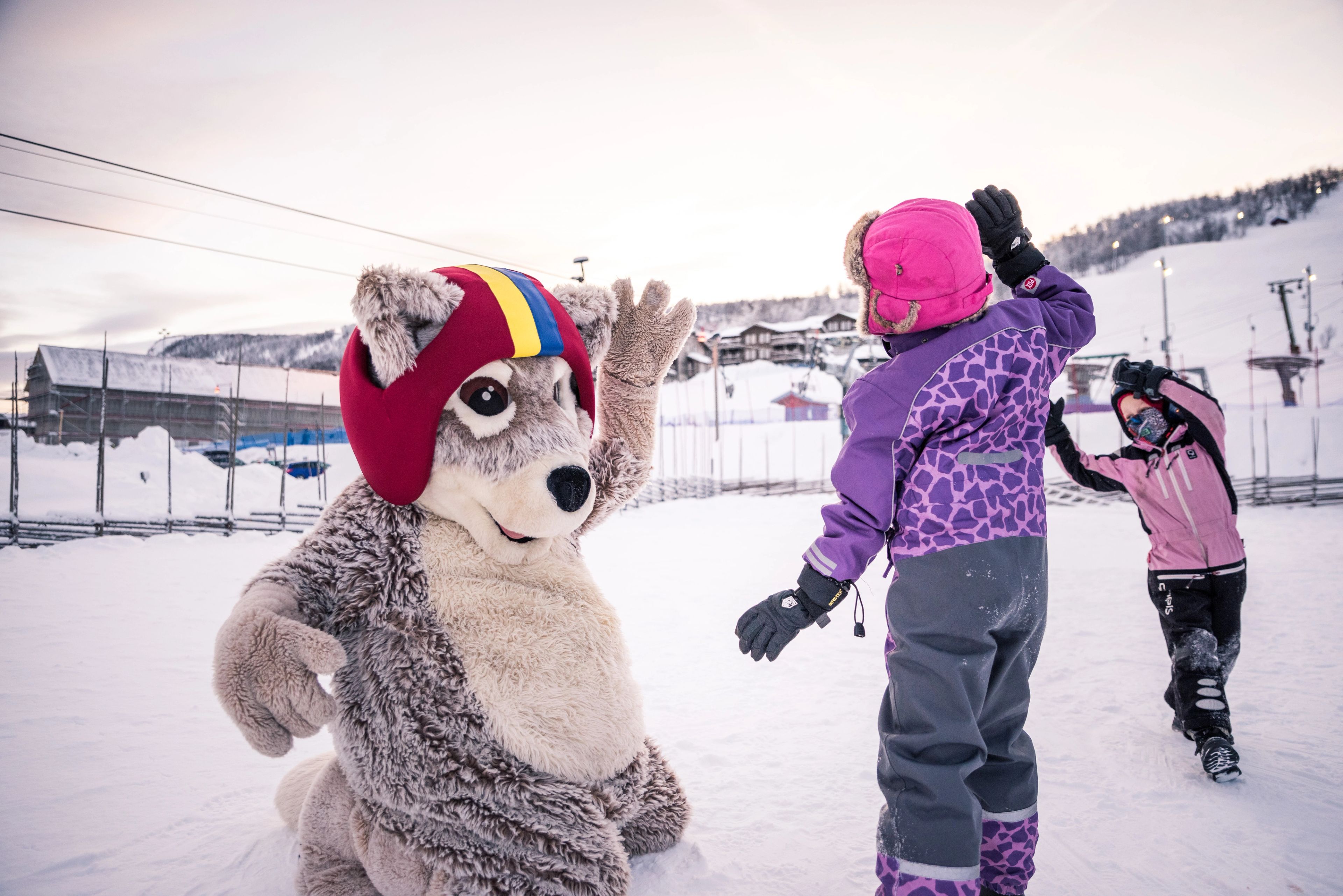 Två barn leker i snön med maskoten Mickelina fjällräv.
