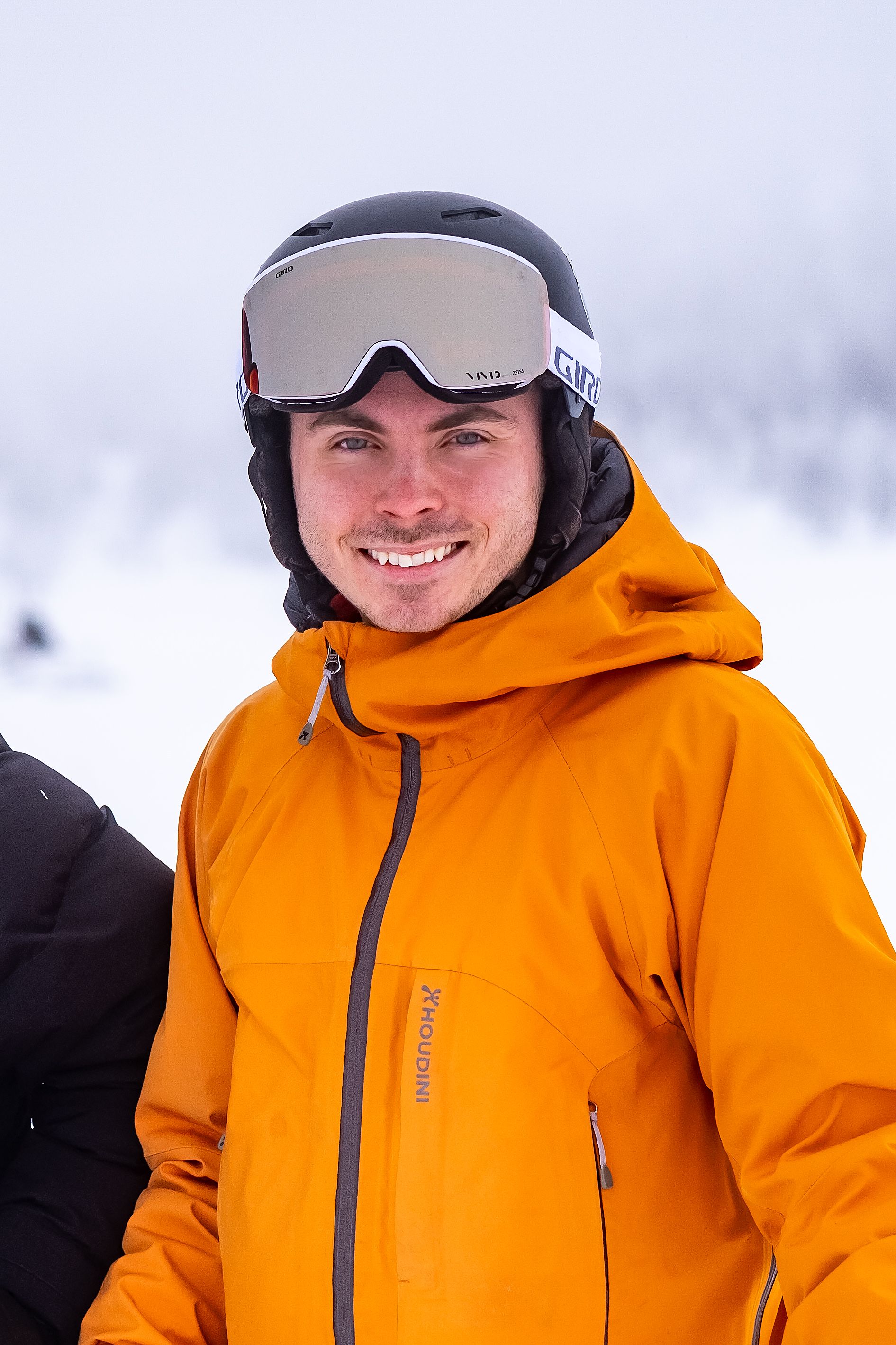 A man in a black ski helmet, reflective goggles, and an orange jacket smiles at the camera on a snowy day.