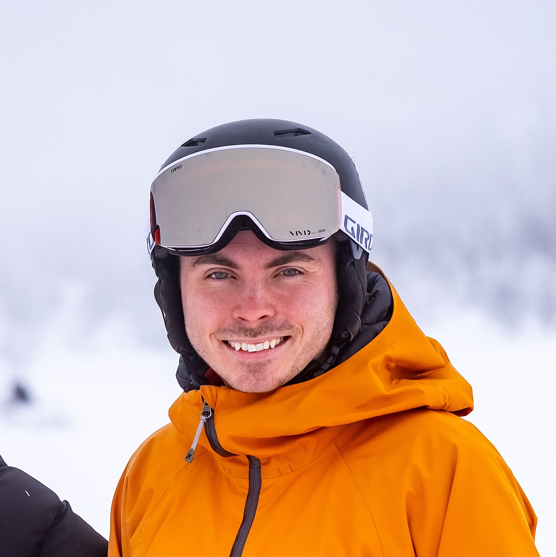 A man in a black ski helmet, reflective goggles, and an orange jacket smiles at the camera on a snowy day.